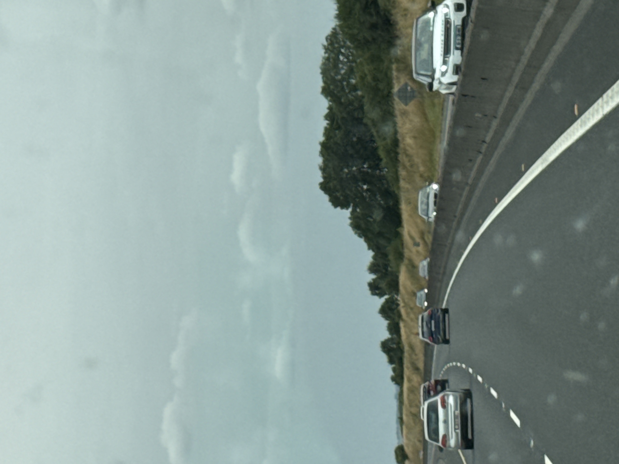 The image shows a highway scene in Pill Belan, Ireland. The road is asphalt with white lane markings. On the road, there are several cars driving in the same direction. In the foreground, a white car is visible on the left. Further ahead, a silver car is in the left lane, followed by a dark-colored car in the same lane. To the right, a white SUV is in the right lane. The environment consists of a concrete barrier separating the lanes, with dry grass and trees visible on the right side of the road. The sky is overcast with clouds. There are no people visible in the image.