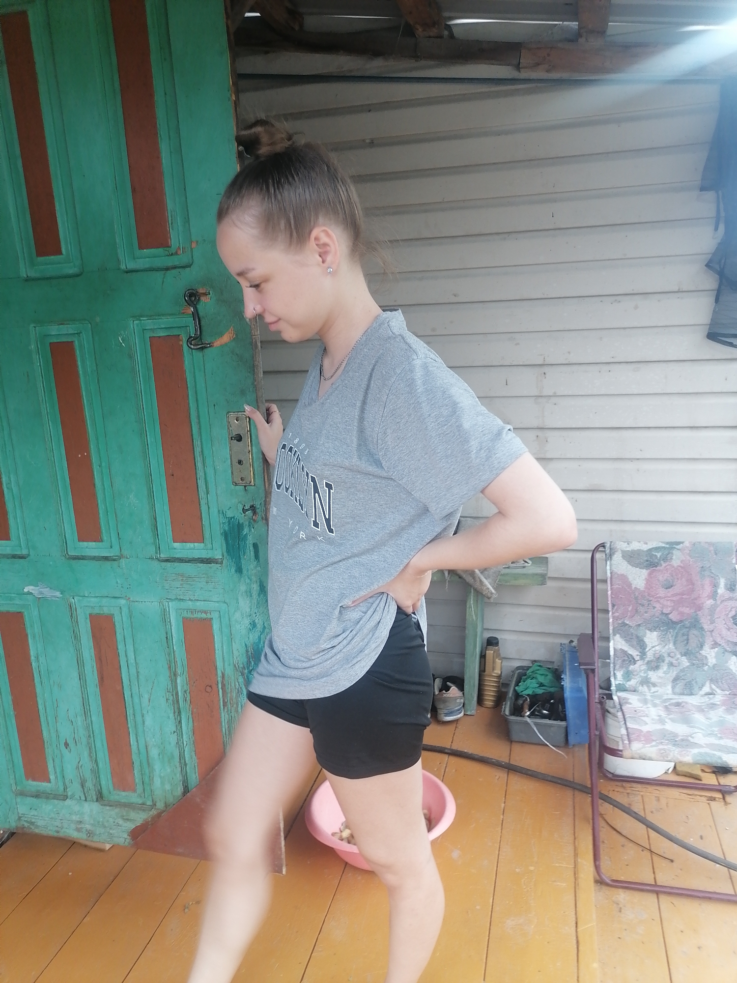 A young woman with blonde hair pulled into a bun stands on a wooden porch. She is wearing a grey t-shirt with the text "BROOKLYN" printed in black and white, and black shorts. Her left hand is on her hip, and her right hand is touching a green wooden door with red accents. The door has a black metal latch. Sunlight streams in from the upper right, creating lens flare.
Behind the woman, the porch wall is made of horizontal white siding. To the right of the door, there is a chair with a floral-patterned cushion. Various objects are on the porch floor, including a pink bowl with what appear to be nuts or seeds, a pair of sneakers, a bottle, and some cleaning supplies. The wooden floor is painted yellow. The scene appears to be outdoors, possibly in a rural or suburban setting, given the rustic door and porch. The time of day is likely daytime due to the bright sunlight. There are no obvious indications of the weather, but the bright light suggests it is clear. The overall mood seems casual and relaxed.