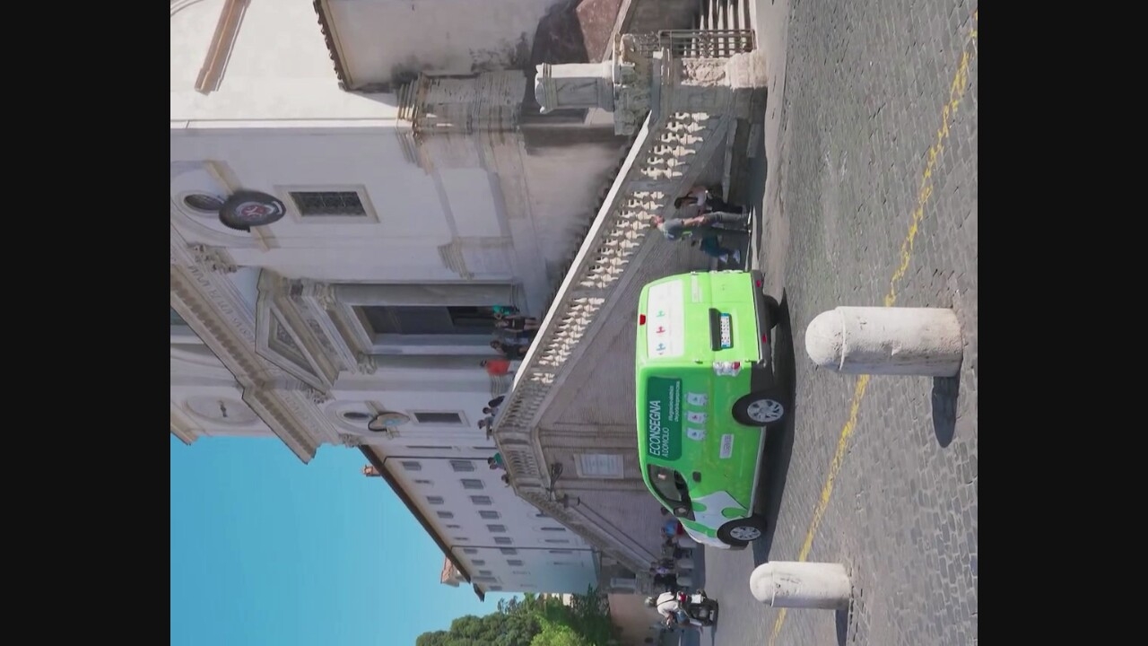 In Rome, Italy, a bright green van with the branding "ECONSEGNA" is parked on a cobblestone street in front of a large, white building with a grand staircase leading to an entrance. The van has white graphics and text on its sides, including images of what appear to be washing machines. Several people are visible in the scene. Some are walking up the stairs, while others are seated on the steps. A person is standing next to the van, and a motorcyclist is seen on the left side of the frame. The building, likely a church or historical monument given its architecture and the presence of people gathered on the steps, is bathed in sunlight under a clear blue sky. Two cylindrical concrete bollards are placed on the street in the foreground.