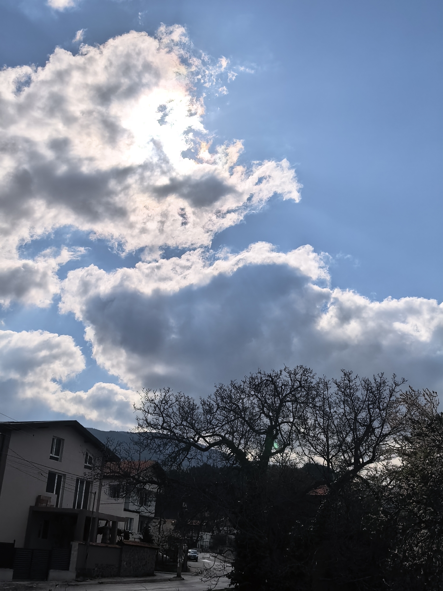 The image presents a daytime outdoor scene in what appears to be a residential area. The sky dominates the upper half of the frame, displaying a bright blue sky with several white and gray clouds, with the sun visible through a gap in the cloud cover. The bottom half of the image includes buildings, trees, and a road. A multi-story building is on the left, with visible windows and a balcony-like structure. Silhouetted trees in the foreground obscure some of the view. A road is visible between the trees and buildings, with a car in the distance. A mountain range is faintly visible in the background. The city and specific location are unidentified.