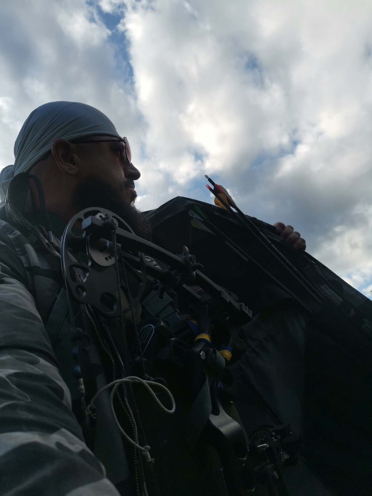A man wearing a bandana and sunglasses is seen from the chest up, looking to the right. He has a beard and is wearing a camouflage jacket. In front of him, a compound bow is visible, along with a quiver holding several arrows. The man is standing outdoors, with a cloudy sky in the background. The image does not provide information about the location in the city of Semypolky, Ukraine, or any specific actions or interactions.