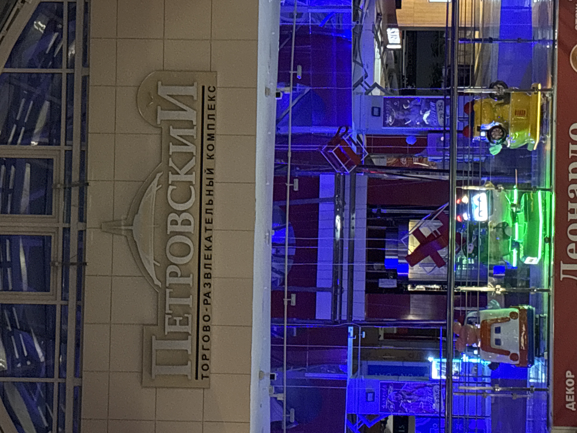 The image shows a partial view of the Petrovsky Shopping and Entertainment Complex in Izhevsk, Russia. The building features a grid-like facade on the left, and a sign with Cyrillic lettering identifying the complex is visible. The interior appears to have a multi-story structure with various attractions or rides, including vehicles in yellow, green, and white/orange. There are also promotional displays featuring posters with Cyrillic text. The lighting in the interior is a mixture of neon blue and other colored lights.