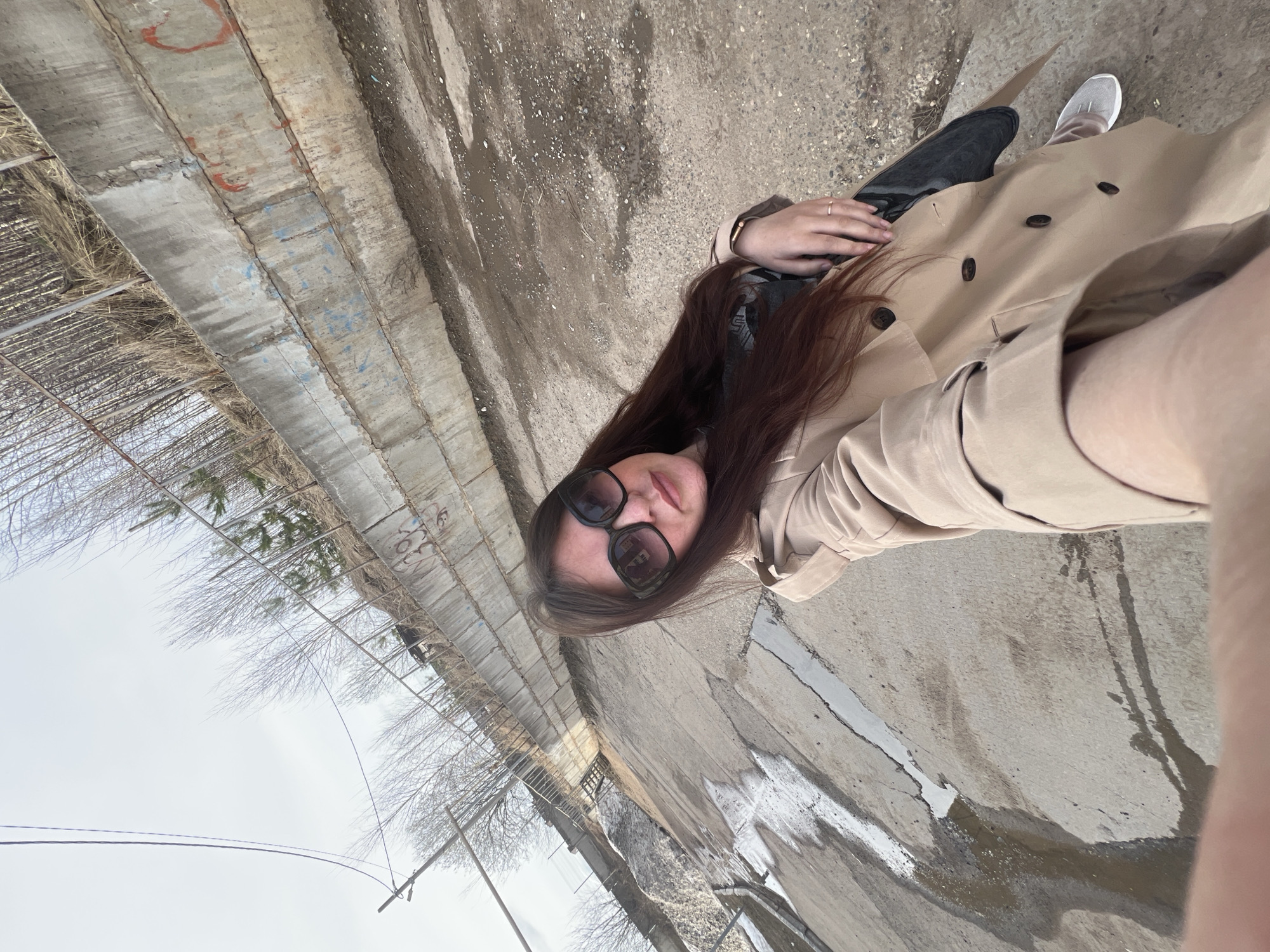 A woman with long brown hair and sunglasses is lying on a concrete surface. She wears a beige trench coat, black jeans, and a light gray shoe. The surface is cracked and stained, possibly from water or other liquids. Beside her, there's a concrete wall with metal fencing and bare trees. The sky is overcast. The location appears to be an outdoor area, likely an urban or industrial zone, somewhere in Noviy, Russia.