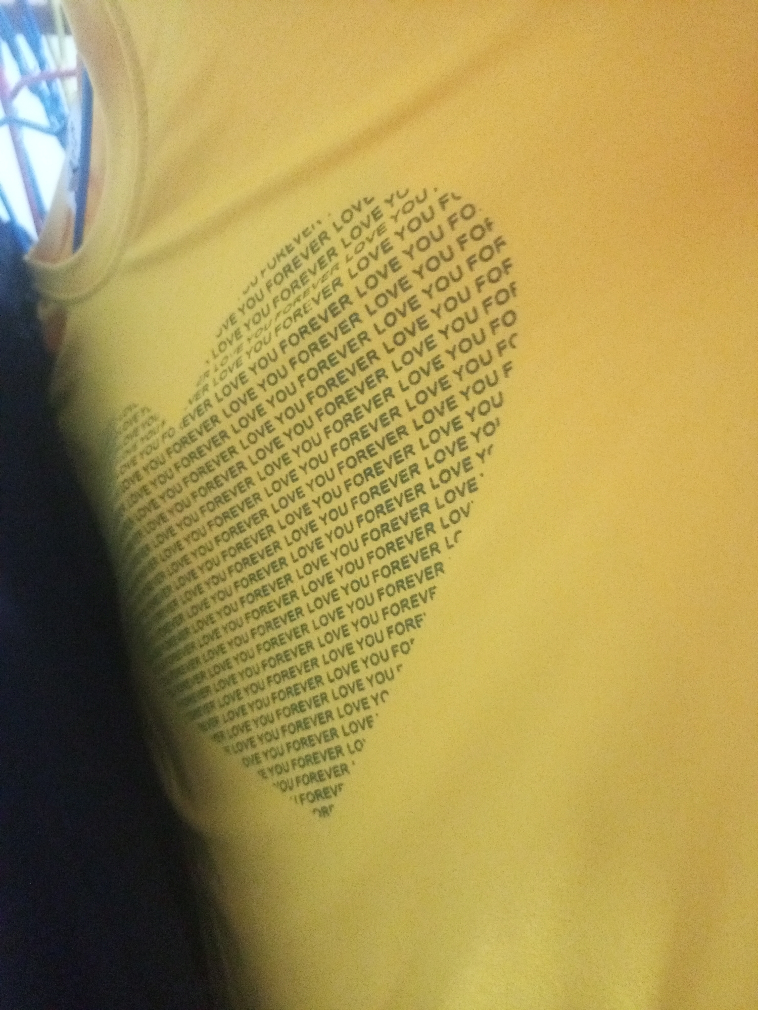 The image is a close-up shot of a yellow garment, possibly a t-shirt, with repeated text printed on it. The text appears to be "LOVE YOU FOREVER LOVE YOU FOREVER LOVE YOU FO" and variations thereof, forming a pattern. The garment is draped and creased, suggesting it is being worn or is folded. A dark, out-of-focus area on the left side of the image likely represents a person's body or shadow. The background is a plain yellow color, consistent with the garment. There is no discernible information about the location being La Tagua, Venezuela.
