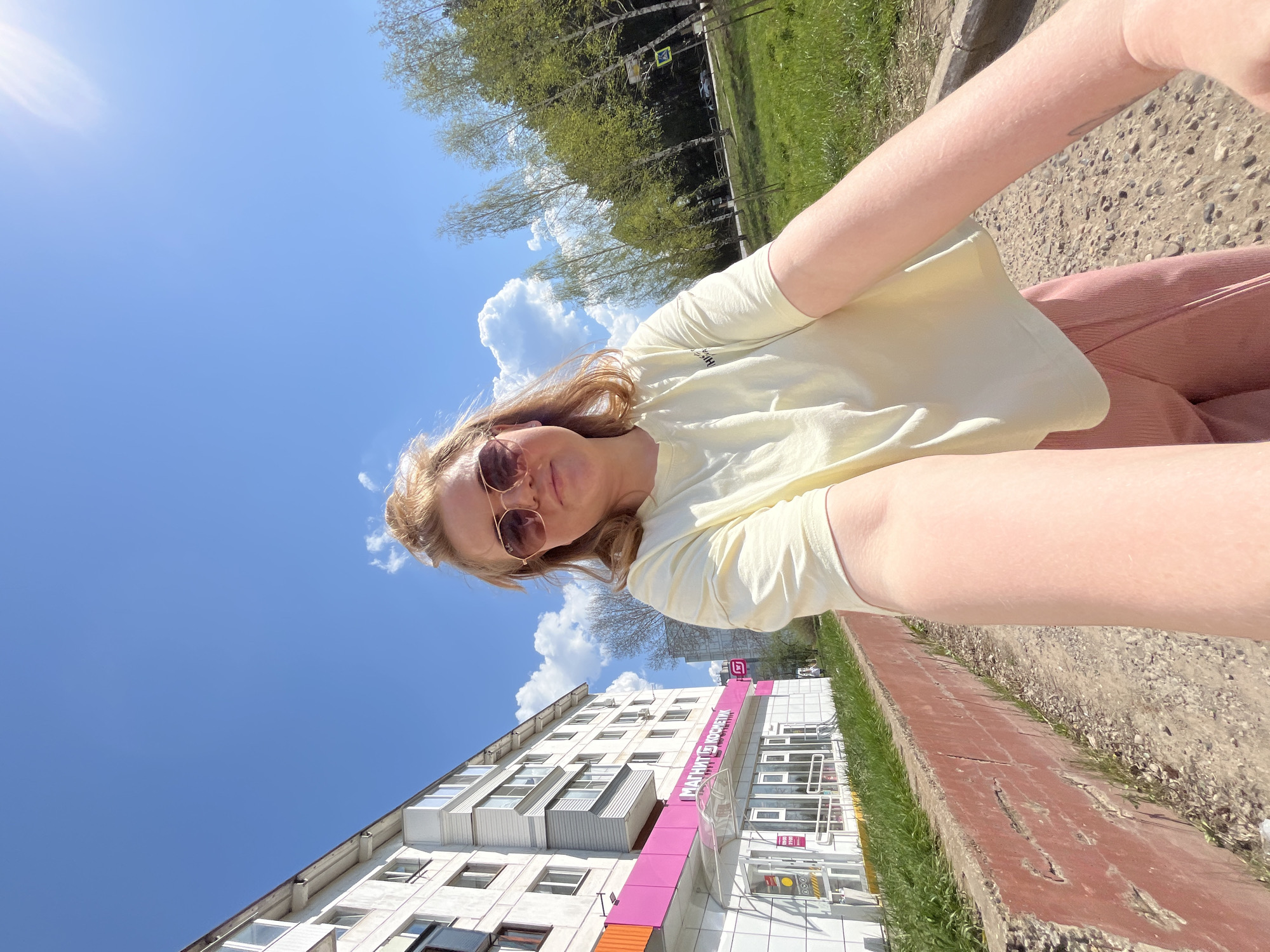 A low-angle, rotated selfie shows a woman with reddish-blonde hair and sunglasses wearing a pale yellow t-shirt and rose-colored pants. She is outdoors on a sunny day with a blue sky and scattered clouds. In the background, a white multi-story building has a storefront with a pink sign displaying Cyrillic text, which identifies it as a branch of the Russian retail chain "Magnit Kosmetik." The photograph was taken in the city of Chaikovsky, Russia. The scene also includes trees and greenery, along with a section of sidewalk.