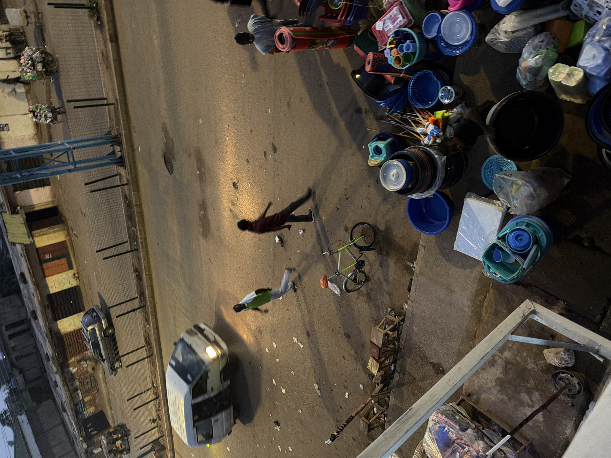 The image depicts a street scene in Bukuru, Nigeria, characterized by a mix of vehicles, pedestrians, and market stalls. A white van is visible in motion, its headlights illuminated. Several people are present, including a person in a green shirt and grey pants walking across the street, and another person in a dark shirt walking in the opposite direction. A bicycle with an orange object on its seat is parked on the side. The ground is a paved road littered with various items, including paper scraps. In the foreground, market stalls are set up with an assortment of goods, primarily plastic containers, bowls, and baskets of varying sizes and colors. Some rolled-up mats are also stacked. The background shows buildings and a blue metal structure. The overall impression is of a bustling, everyday street environment, possibly in the early morning or late evening, given the lighting conditions and the presence of illuminated headlights.
