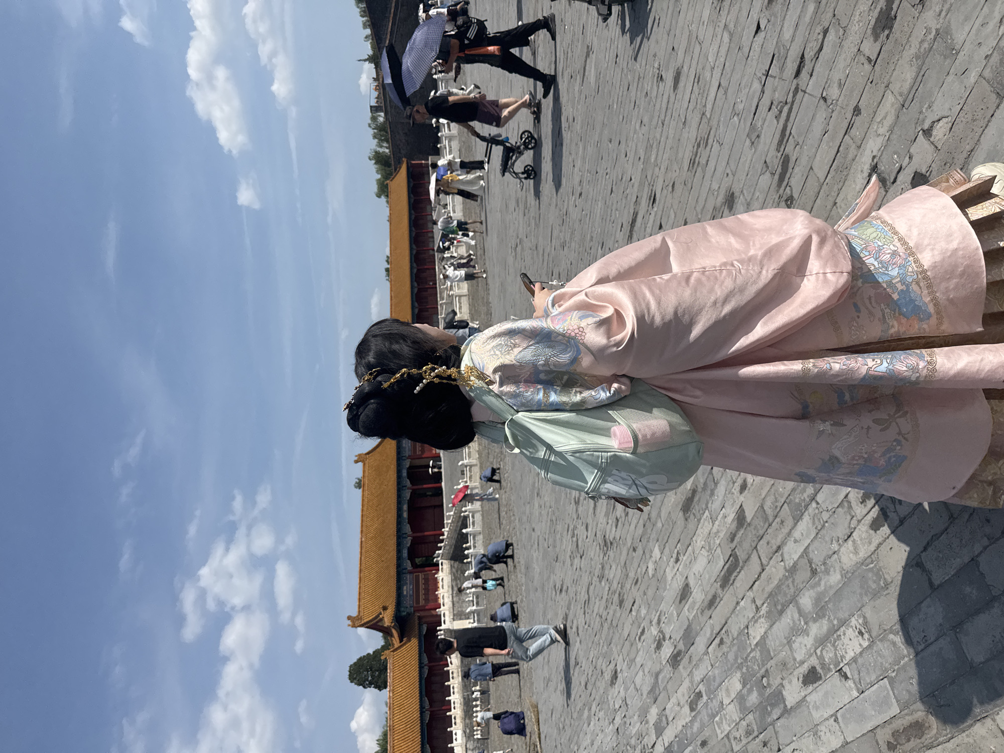 In the city of Dongcheng District, China, a young woman stands in the foreground, facing away from the camera and looking to her right. She is wearing a traditional light pink Hanfu dress adorned with intricate patterns and a mint green backpack. Her hair is styled in an elaborate updo with gold ornaments. She holds a smartphone in her right hand. Behind her, a large, paved courtyard stretches out towards a historic palace complex with orange-tiled roofs and red walls. Several other people are scattered throughout the courtyard, some walking, others sitting, and a few are holding umbrellas to shield themselves from the sun. The sky above is blue with scattered white clouds. The ground is paved with grey bricks.