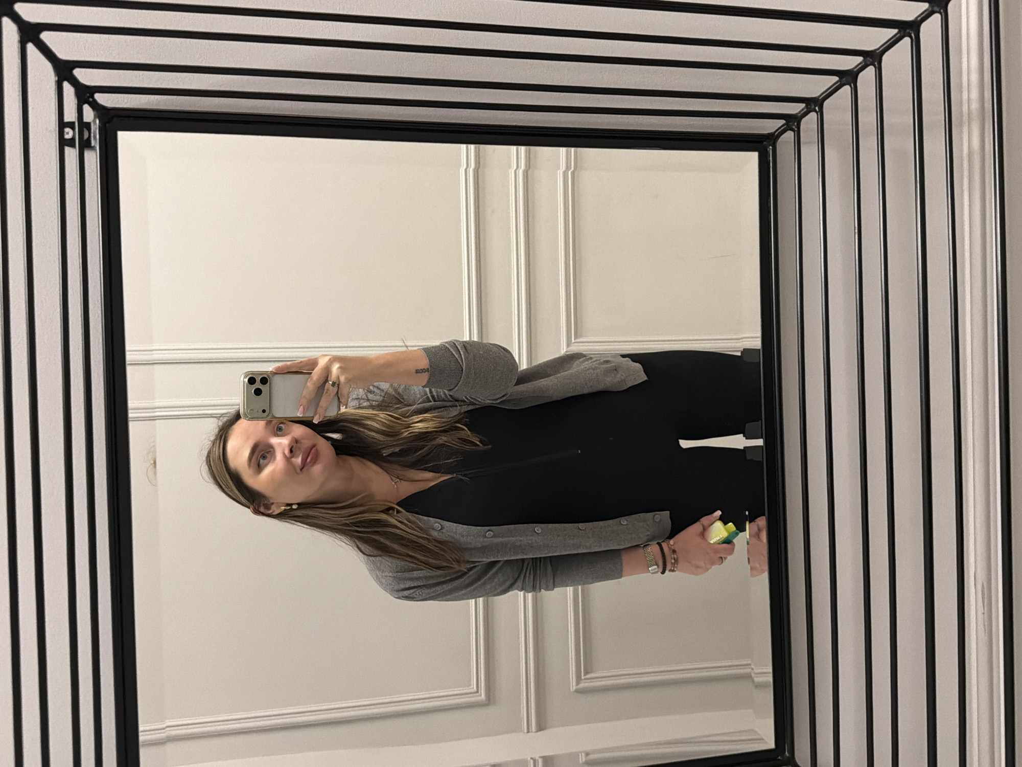 A full-body mirror selfie shows a woman holding a smartphone, capturing her reflection in an ornate black-framed mirror. The mirror features a distinctive geometric grid frame made of thin black bars, creating a complex pattern around the reflective surface.
The woman has long, light brown hair with highlights, styled down, and is looking slightly upwards towards her phone with a subtle smile. She is wearing a light grey button-up cardigan over a black top or dress. On her left wrist, she wears a watch with a metallic band and several beaded bracelets. On her right hand, she holds a small, light green and yellow object, possibly a makeup sponge or a small container. A small tattoo with the numbers "2004" is visible on her left forearm.
The background reflected in the mirror consists of light-colored walls, likely white or off-white, adorned with decorative molding or wainscoting, suggesting an interior space like a room or hallway. The scene appears to be well-lit indoors. The overall impression is a casual, everyday moment captured in a stylish setting, consistent with an indoor location in Moscow, Russia.