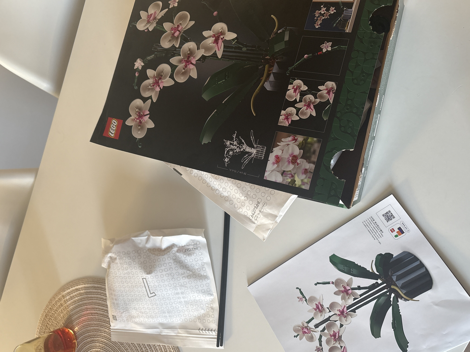 The image displays an overhead view of a white table, set up for assembling a LEGO Orchid botanical set. The primary subject is the open box of the LEGO Orchid set, featuring a large image of the intricate brick-built orchid with white and pink flowers and green foliage. Below this, an outline drawing indicates the set's dimensions as "39 cm / 15.4 in.", and the red LEGO logo is visible.
Alongside the box, a white instruction manual for the LEGO Orchid is spread out, showing a rendered image of the assembled orchid in a dark pot. The manual's cover includes multilingual text indicating availability of the booklet in English, German, French, Italian, and Spanish, as well as a note for a Chinese version and a QR code with "QLEGO® Builder."
Two white plastic bags filled with LEGO elements are also present. One visible bag is labeled with the number "6427369" and a large letter "L", while another partially obscured bag shows "6427367". A clear glass containing an amber liquid, resting on a round, woven placemat, occupies the lower-left corner of the frame. The setting appears to be indoors during the daytime, brightly lit. The scene suggests a relaxed activity taking place in Santiago de Compostela, Spain.