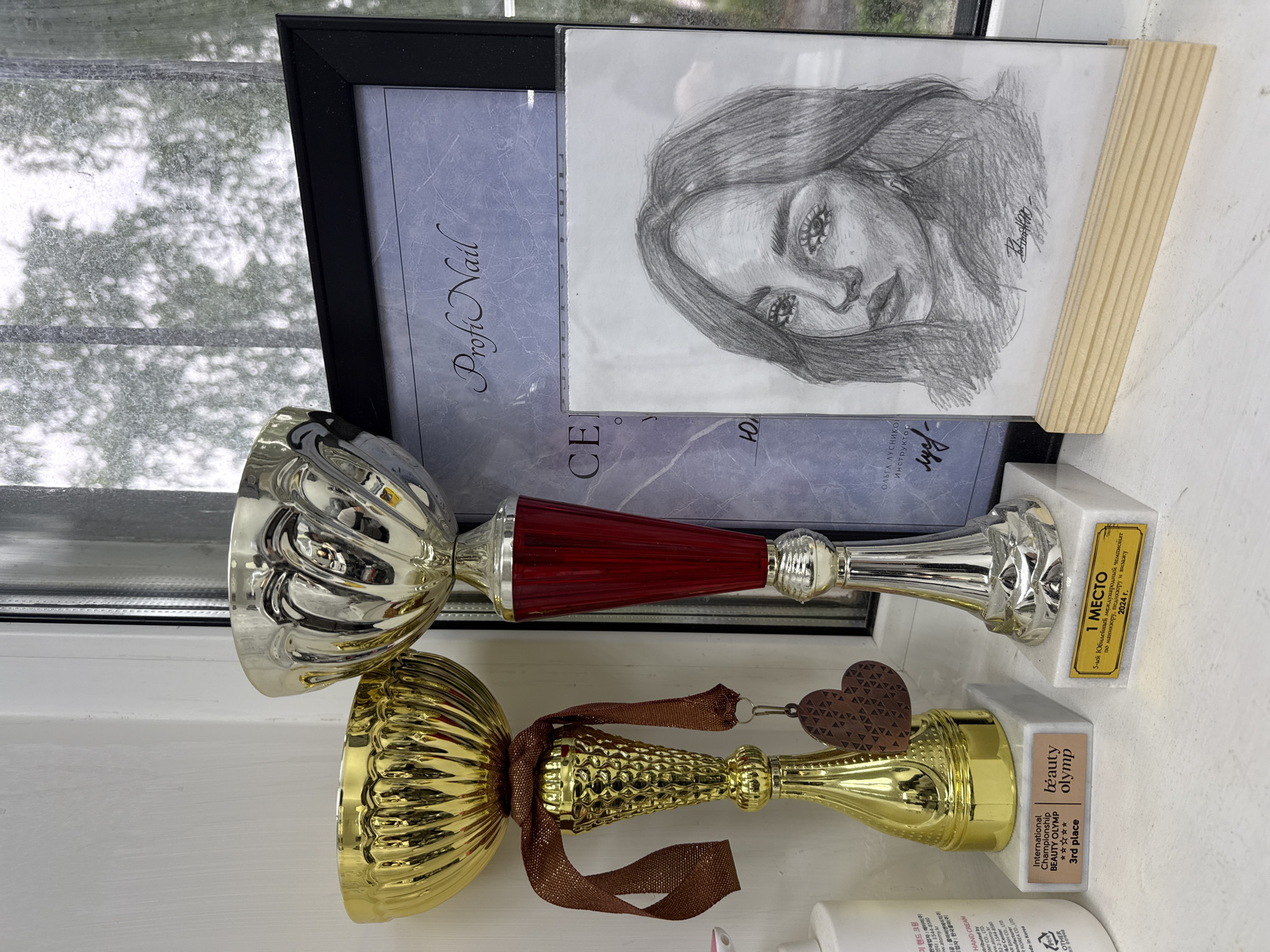 The image displays a collection of awards and personal items arranged on a white windowsill, set against a window revealing a blurry, light-filled outdoor view, possibly trees or foliage.
**Subjects:** The primary subjects are two trophies, a framed certificate, a pencil drawing, and a partially visible cosmetic bottle.
**Objects:**
* A taller, slender silver trophy with a distinctive red ribbed stem, perched on a white marble-like base.
* A shorter, ornate gold trophy with a textured surface, adorned with a brown ribbon and a heart-shaped charm, also on a white marble-like base.
* Behind the trophies, a black-framed document or certificate features elegant script, partially reading "Profi Nail."
* Leaning against the framed document is a detailed pencil sketch of a woman's face, notable for its expressive eyes and dark hair, with a visible signature.
* In the lower right corner, a white bottle, labeled in Korean and English as "HAND CREAM" from "ATOMY CO., LTD" (Made in Korea), is partially visible.
**Activity or Event:** The items collectively represent achievements in a professional or competitive context, specifically within the beauty industry. The trophies and certificate signify awards for excellence.
**Notable Details:**
* The silver trophy's base proudly displays "1 МЕСТО" (1st place) and details "5-ый Юбилейный международный чемпионат по маникюру, педикюру и визажу" (5th Anniversary International Championship in manicure, pedicure, and makeup) for "2024 г." (2024 year).
* The gold trophy's base reads "International Championship BEAUTY OLYMP" and "3rd place," along with five stars.
* The framed certificate mentions "ОЛЬГА ЛУСНИКО Инструктор" (Olga Lusniko Instructor), indicating the recipient's role.
* The setting is likely a home or studio in Chaikovsky, Russia, given the Cyrillic text on the awards. The natural light suggests a daytime setting. The drawing reflects a personal artistic touch.