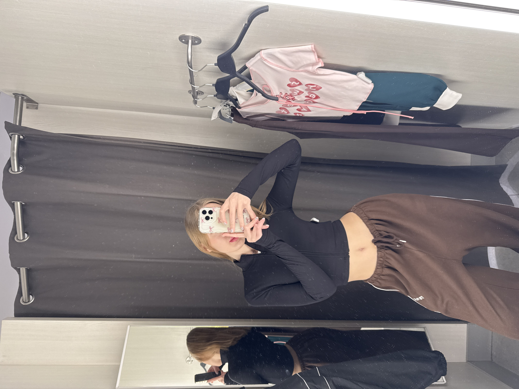 The scene depicts a female subject taking a mirror selfie inside an indoor changing room, located in the city of Rostov-on-Don, Russia. The subject, a woman with long, light brown hair, is positioned centrally, partially obscuring her face with a smartphone held in both hands. She wears a black, long-sleeved, cropped athletic top with a front zipper and loose-fitting brown sweatpants featuring a drawstring waist and white side stripes; her navel is exposed. The smartphone is equipped with a clear, patterned case.
To the left, a full-length mirror reflects the subject's back, showing the back of her black top and brown sweatpants. The changing room is enclosed by a dark grey privacy curtain on a silver rod, visible behind the subject. The walls are a light, textured off-white. To the right, a silver metal clothing rail is mounted on the wall, holding several items of clothing on hangers. These include a light pink short-sleeved top with a lace-up front and multiple red heart designs, a dark brown garment, a white garment, and a dark green garment with a white cuff. The lighting is uniform, suggesting overhead illumination. No other individuals or external identifiers are visible.