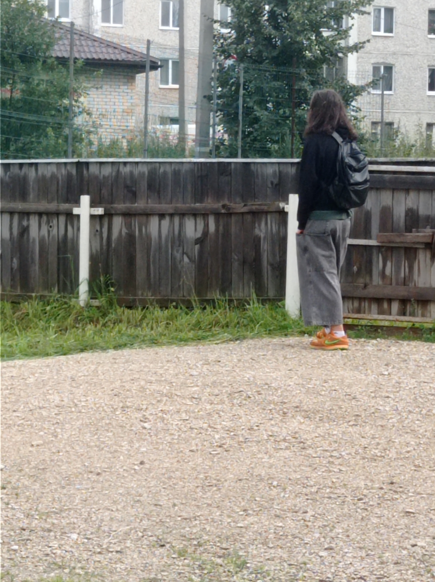 The image shows a person from the back, standing on a gravel surface in front of a wooden fence. The person is wearing a black jacket, grey wide-leg pants, and orange sneakers. They have a black backpack on. Behind the fence, there are trees and a multi-story building with windows. To the left of the person, a white cross-shaped object is attached to the fence. The scene appears to be an outdoor area in the city of Degtyarsk, Russia, possibly a residential backyard or a common area in a housing complex.