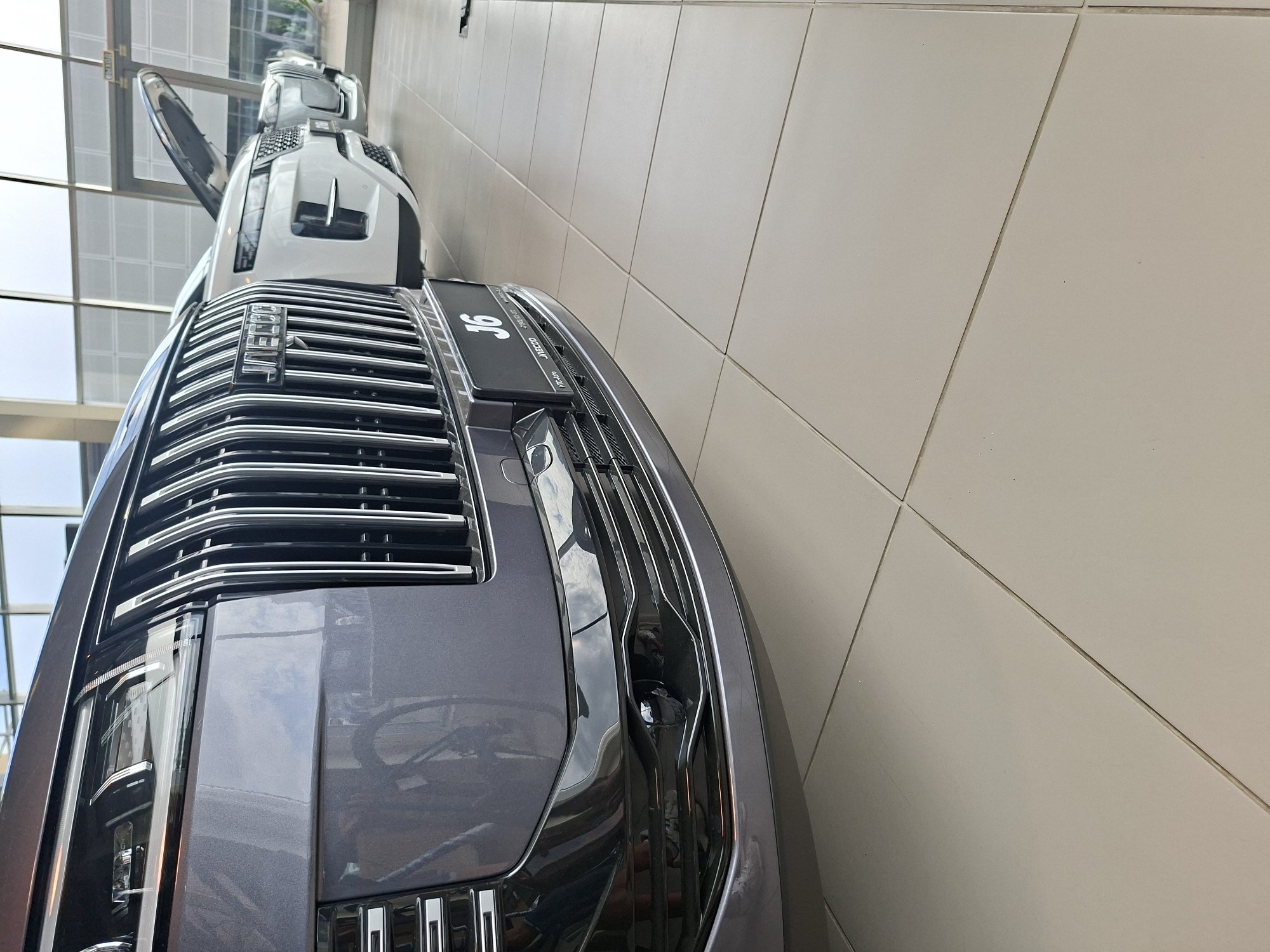 In Izhevsk, Russia, a close-up shot showcases the front of a dark gray SUV parked on a light-colored tiled floor inside a modern showroom. The vehicle's prominent chrome grille features vertical slats and a "JAECOO" logo. Below the grille, a black license plate holder displays "J6" and other text. The car's headlight is visible on the left, and a section of a white SUV with its hood open, and another vehicle are parked behind it. The background shows large windows and architectural elements of the building.