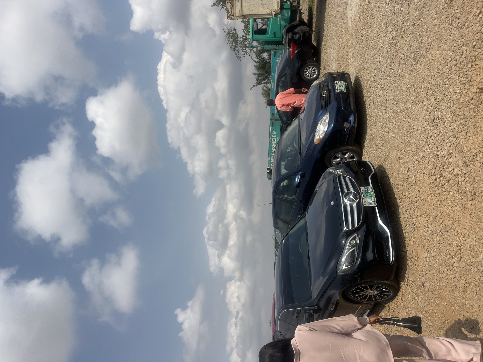 The image depicts a partially overcast day in what appears to be a parking area in Galadimawa Village, Nigeria. Several vehicles are present, including a black Mercedes-Benz with license plate AGL726GK visible in the foreground, followed by a dark blue Toyota, and a green truck in the background. A person in a peach suit is visible next to the Mercedes, holding a black object. Further back, another person is standing by a black car. The ground surface is covered in gravel.