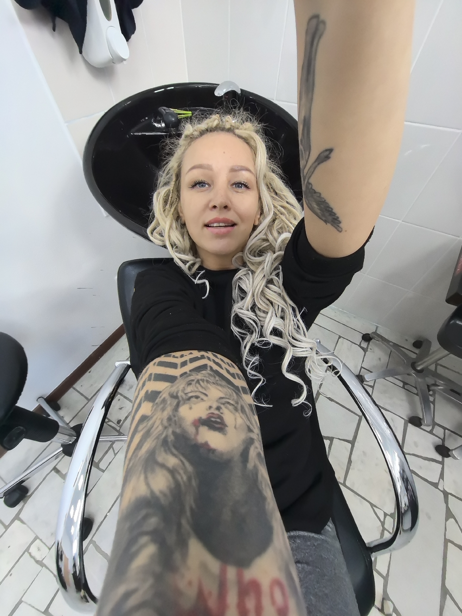 An overhead shot captures a woman with blonde, braided hair sitting in a salon chair with a black basin behind her. She is wearing a black long-sleeved shirt and gray pants. Her right arm is extended toward the camera, revealing a tattoo of a woman's face. Another tattoo of what appears to be a bird in flight is visible on her left arm. The floor is tiled with a light-colored, geometric pattern. The scene suggests a hair salon or similar establishment. The location is the city of Chaikovsky, Russia.