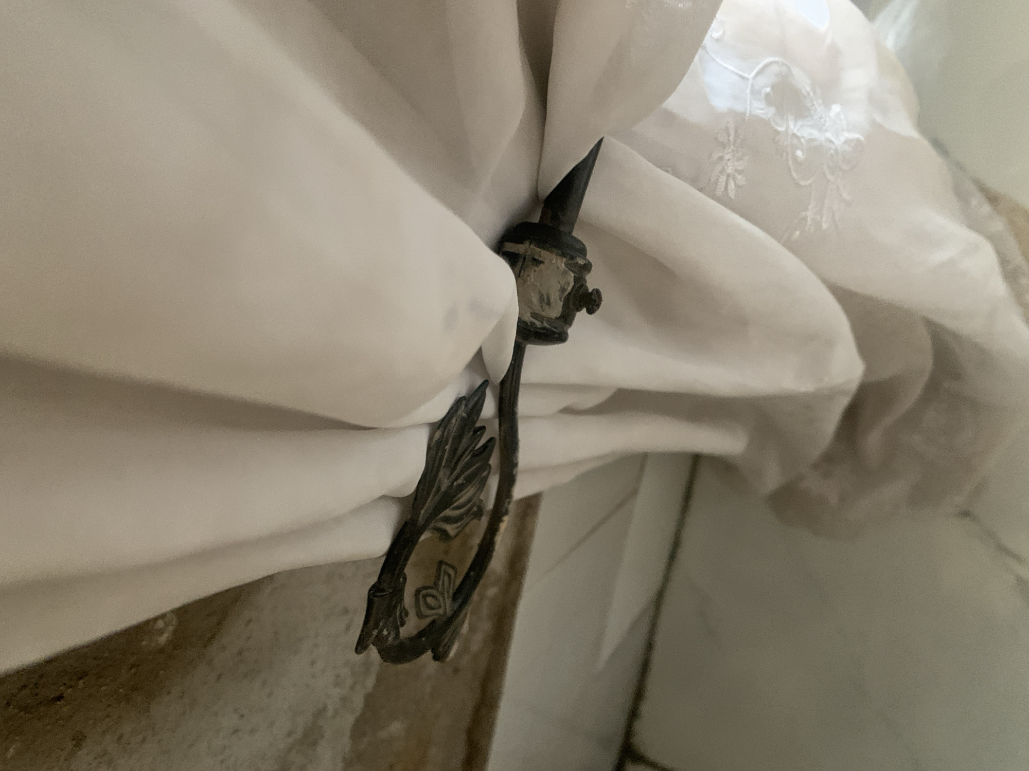 A black metal curtain tie-back shaped like a bird's wing is securing a white sheer curtain. The tie-back is attached to the wall behind the curtain. The wall appears to be textured and possibly stained. Below the curtain, a white tiled floor is visible. The scene suggests an indoor setting, likely a room with a window.