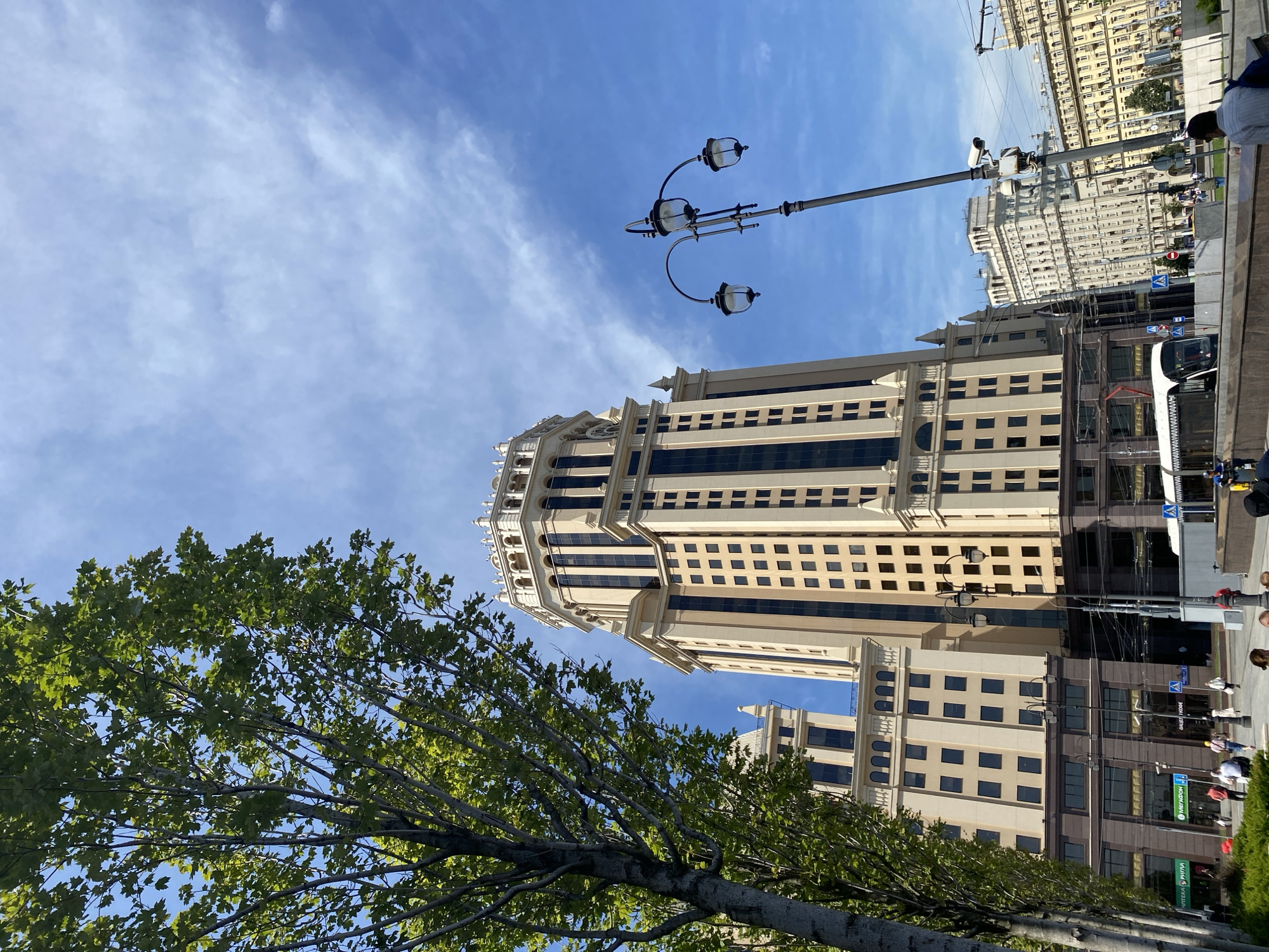 The image shows a street view in Moscow, Russia, featuring the prominent Stalinist skyscraper, the Ministry of Foreign Affairs building, located at Smolenskaya Square. In the foreground, a mature tree with green foliage partially obscures the view of the building. A lamppost with three lights stands to the right of the building, and in the background, other buildings and a tram are visible. The sky is blue with scattered white clouds. Several people are seen walking on the sidewalk and crossing the street, engaged in everyday activities.