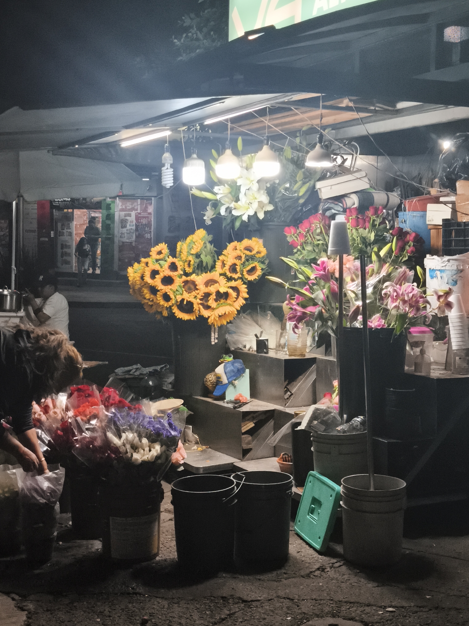 This is an outdoor nighttime scene in Mexico City, Mexico, depicting a flower stand under a canopy lit by several hanging lights. The stand displays an assortment of flowers including sunflowers, lilies, and roses, arranged in buckets and vases. A person on the left side of the frame is arranging or selecting flowers. In the background, other individuals are visible entering or exiting a brightly lit storefront across the street. The ground is cracked pavement with buckets scattered around the flower stand.