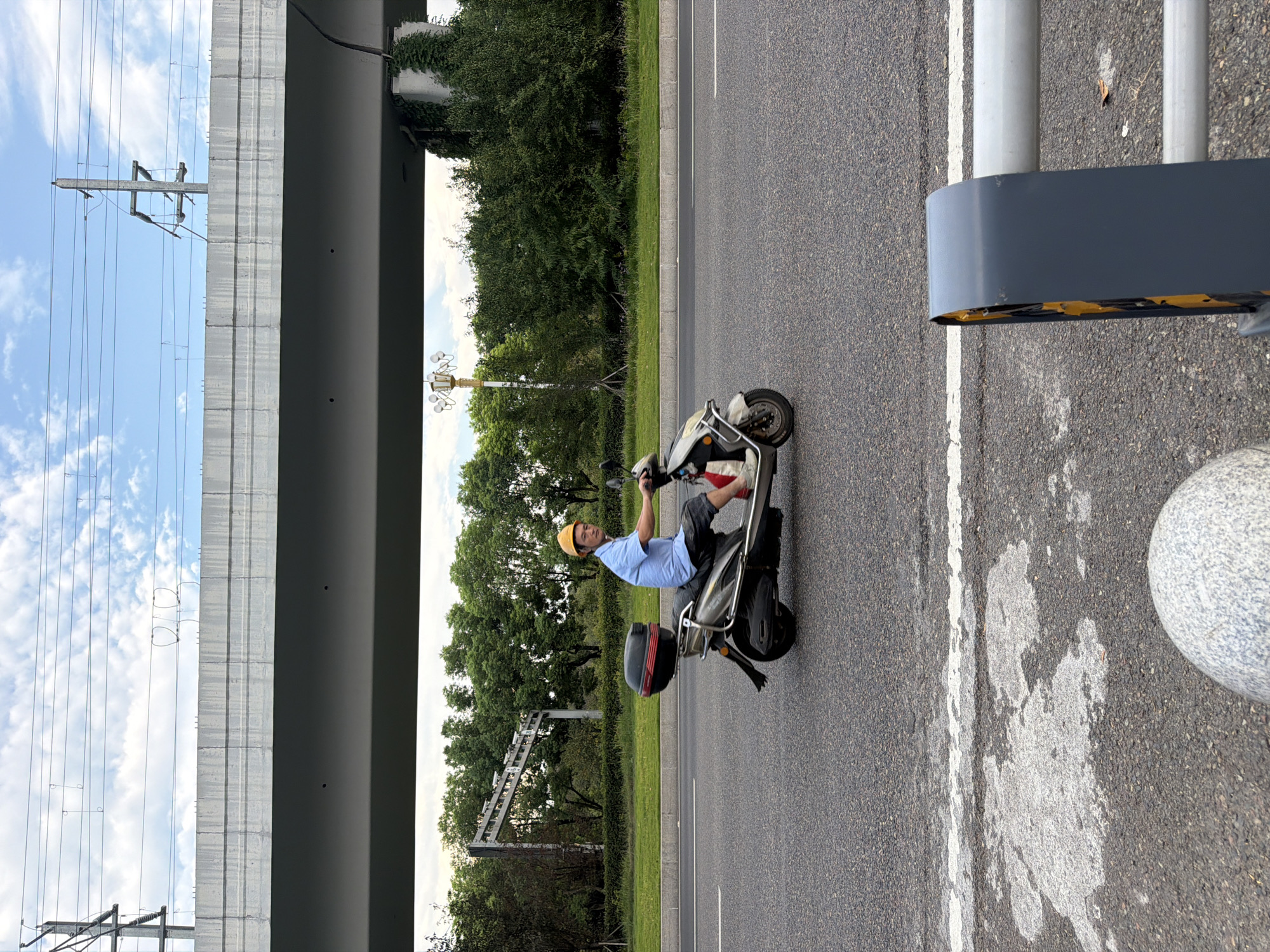 A man wearing a blue shirt, dark shorts, and a yellow hard hat rides a silver and black electric scooter on a paved road. The scooter has a black storage box on the back. The man is looking towards the camera. The road is a multi-lane highway with white lane markings. In the background, there is a large concrete overpass, trees, and power lines. The sky is cloudy. The foreground shows a section of the road with some white paint markings and a gray metallic barrier. The location is identified as Jiangjin, China.