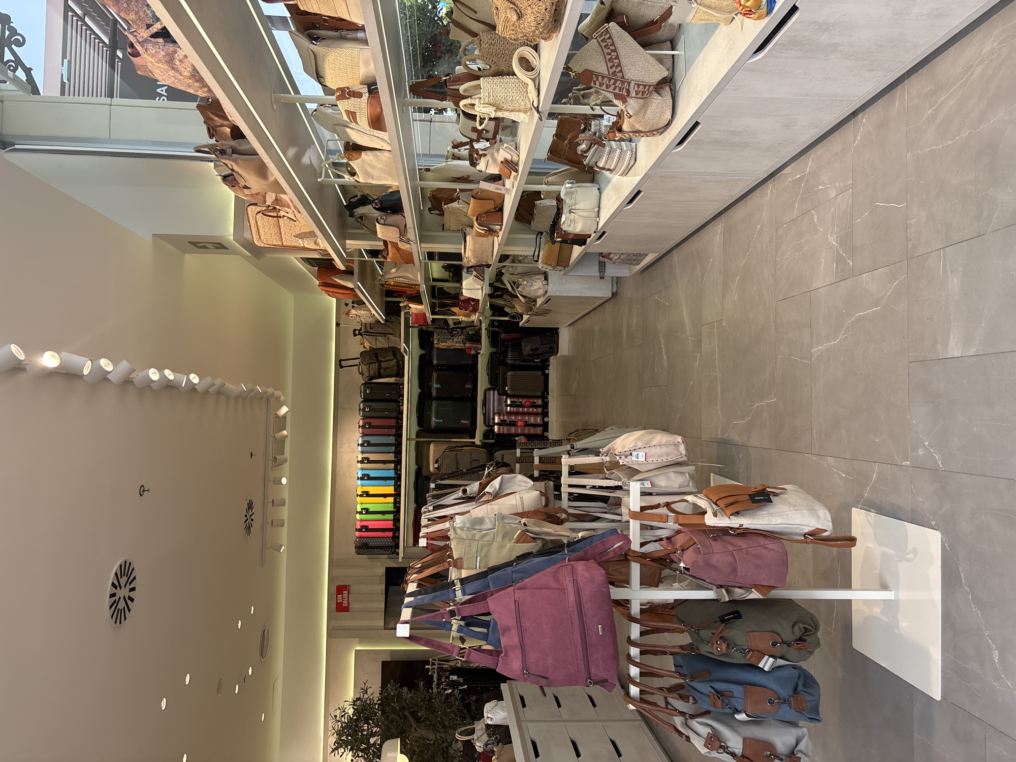 The image depicts the interior of a retail store, likely a handbag and luggage shop, situated in the city of Orihuela, Spain. The store is well-lit with a combination of ceiling lights and track lighting, illuminating rows of merchandise displayed on shelves and racks. The foreground features a white display stand with several handbags of various colors and styles hanging from it. Beyond this, more handbags are visible on shelves lining the walls. In the background, a wall displays a row of colorful suitcases. The flooring is a light gray tile with a marble-like pattern. The overall ambiance suggests a clean, organized, and modern retail environment. There are no people visible in the image. The store appears to be empty, possibly before or after operating hours. The signage visible is in Spanish, indicating the location.