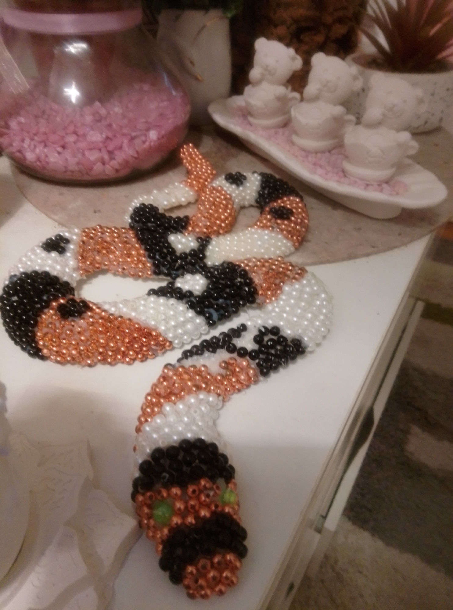 The image shows a beaded snake craft lying on a white surface. The snake is coiled and appears to be made of copper, white, and black beads. The head of the snake is visible at the bottom right, with green beads for eyes and a black beaded mouth. In the background, there is a clear glass jar filled with pink pebbles, and a white tray with small white figurines of bears and cups. The scene is indoors, possibly in a home, and no specific location in Wedel, Germany can be identified from the image.