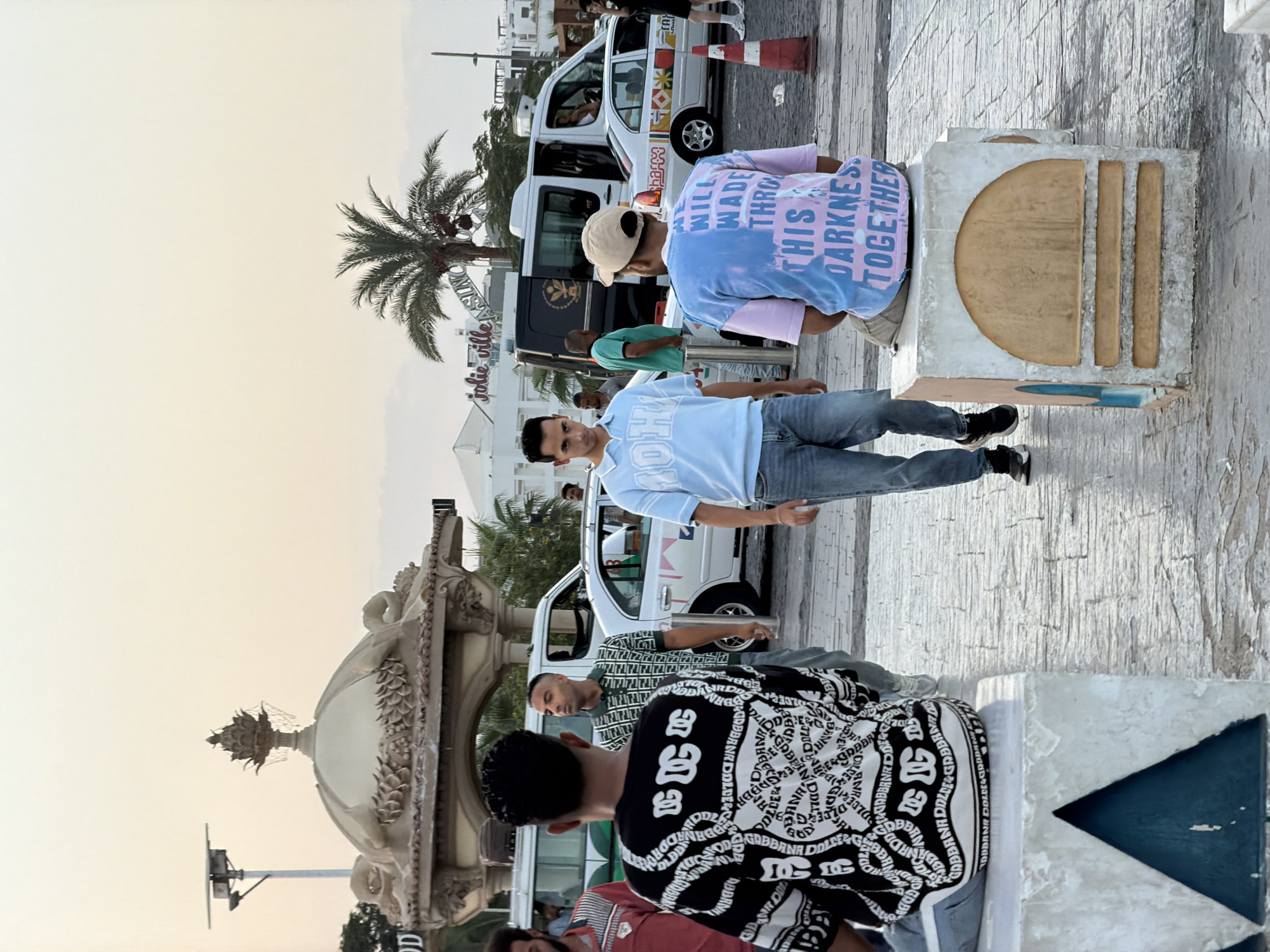 In Sharm El Sheikh, Egypt, a group of men stand in what appears to be a public plaza or street. The time of day is late afternoon or early evening, indicated by the soft, warm lighting and the pale sky.
In the foreground, two men are seated on concrete structures that serve as benches. The man on the left, seen from behind, wears a black and white patterned long-sleeved shirt. The man on the right is also seen from behind, wearing a light pink and blue tie-dye t-shirt with text that reads, "WE WILL WADE THROUGH THIS DARKNESS TOGETHER". He is wearing a tan baseball cap.
To the left and slightly behind the seated men, a man in a black and white patterned shirt stands facing away from the camera. To his right, a man wearing a green and white patterned polo shirt is visible. Further back, a man in a light blue collared shirt walks away from the camera, and another man in a green t-shirt stands near a van.
Several vehicles are present, including white vans and a white sedan. In the background, a distinctive ornate dome structure with decorative carvings is visible, along with palm trees and buildings, suggesting a resort or entertainment area. A red and white traffic cone is also positioned on the ground. The ground is paved with stone tiles. The overall scene depicts a casual outdoor gathering.