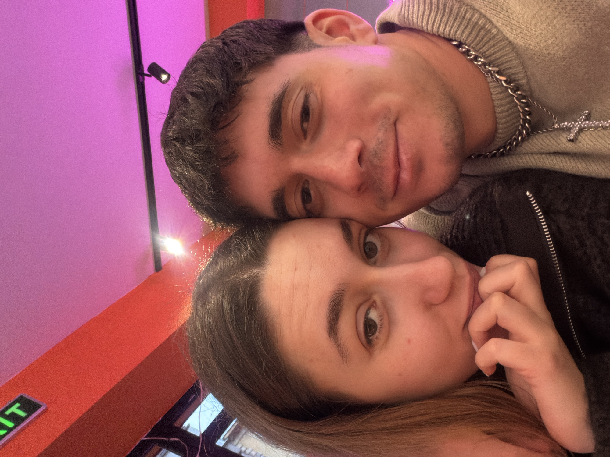 A selfie photo depicts a man and a woman close together. The man has short, dark hair, light skin, and a silver chain with a cross pendant. He is wearing a light-colored sweater. The woman has long, light brown hair, light skin, and is holding her hand near her mouth. The background features a pink wall with a spotlight and an orange surface with a green “EXIT” sign. The location appears to be indoors. There is no evidence to confirm this photo was taken in Sofia, Bulgaria.