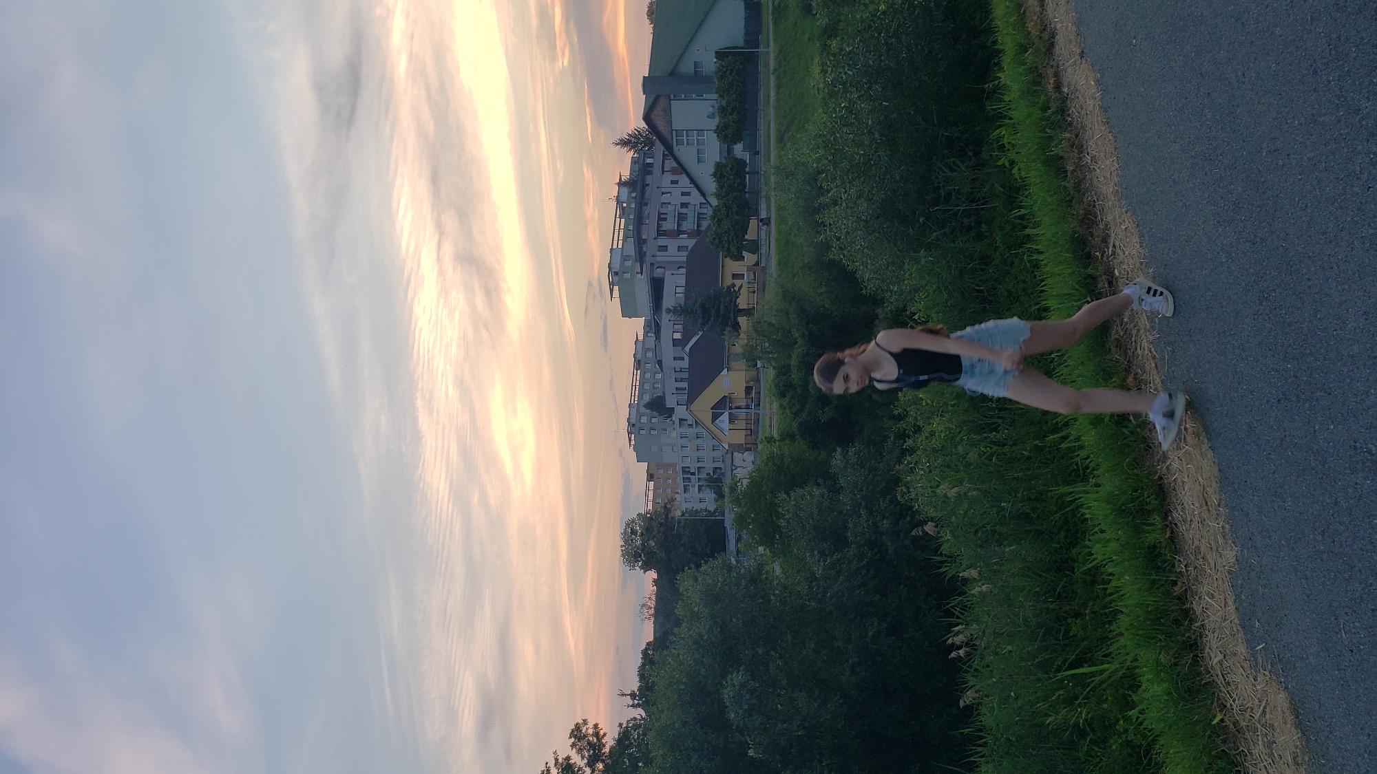 The image depicts a young woman walking on a paved path in Prešov, Slovakia. She is wearing a black tank top and light blue denim shorts. Her right leg is extended forward, and her left leg is bent at the knee, indicating movement. She is wearing white sneakers with black accents. To her left, there is a dense area of green grass and vegetation, with a faint hint of a body of water or a ditch. In the background, there are trees and buildings. The buildings are varied in height and style, with some appearing to be apartment complexes and others smaller structures. The sky above is a pale blue with wispy clouds, tinged with the warm colors of sunset or sunrise, suggesting it is either early morning or late evening. The overall scene is tranquil and suggests an outdoor recreational activity.