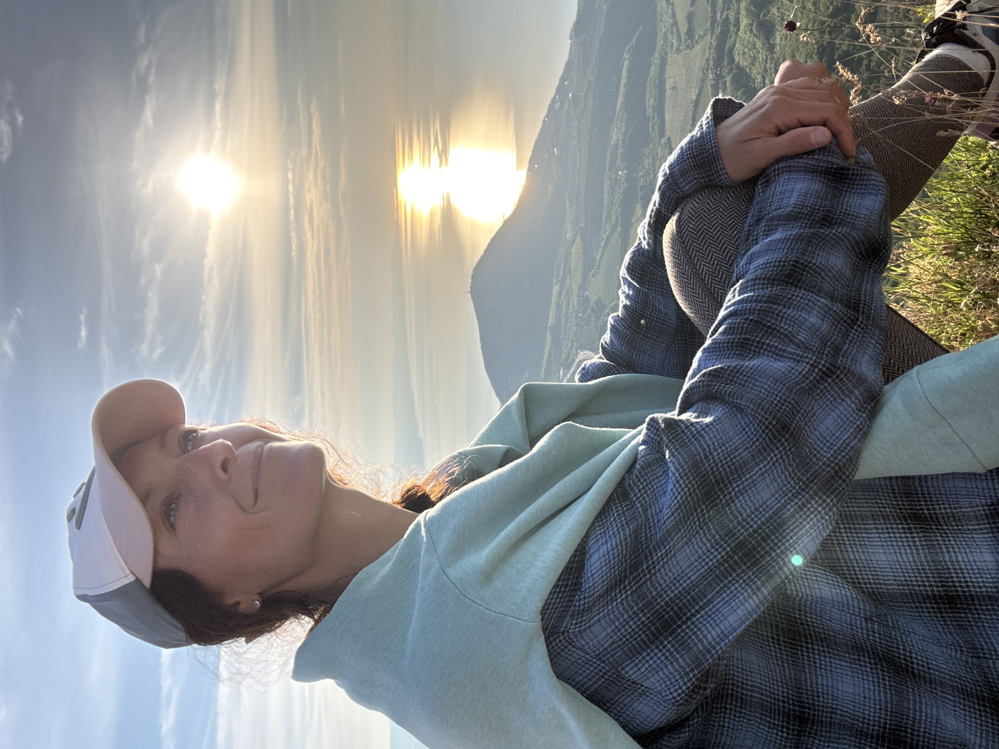 An adult female is seated on a grassy hillside, looking towards a setting sun over a large body of water. The woman, appearing to be in her 30s or 40s, wears a white baseball cap with a light grey brim and an unknown grey emblem, a light blue and dark grey plaid long-sleeved shirt, and a mint green hooded sweatshirt draped over her shoulders. Her hair, dark brown, is pulled back, with some strands catching the warm light. She has a slight smile and is looking towards the right side of the frame, where the sun is low on the horizon, creating a bright reflection on the water. Her hands are clasped around her bent knees, revealing patterned brown and white leggings and dark athletic shoes with white soles. The foreground consists of sunlit green and dry grass with a few small purple wildflowers.
The background features an expansive landscape under a pale blue sky with wispy clouds. A prominent, conical mountain or large hill is visible in the mid-ground, extending into the calm body of water, which is likely a sea or a large lake. Beyond this, further distant landmasses are visible on the horizon, softened by atmospheric haze. The sun, a brilliant white-yellow orb, casts a strong, elongated vertical reflection across the water's surface. Below the mountain, the terrain consists of rolling hills covered in green vegetation, possibly cultivated fields or vineyards, interspersed with forested areas. The lighting is characteristic of either sunrise or sunset, casting long shadows and illuminating the scene with a golden hue. The image is captured in an outdoor environment, likely a high vantage point overlooking a coastal or lake region, in the city of Unknown, Unknown.