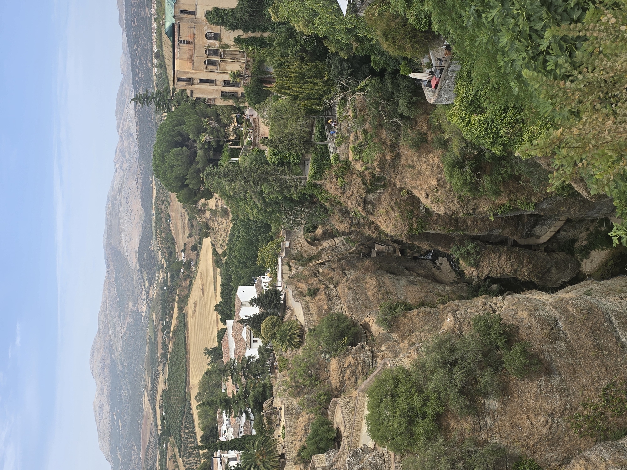 This is a high-angle, wide shot of the El Tajo gorge and Puente Nuevo bridge in Ronda, Spain. The gorge is a deep chasm with sheer, rocky sides that are partially covered with vegetation. The Puente Nuevo, a stone arch bridge, spans the gorge, connecting the old and new towns of Ronda. Buildings and terraced gardens are visible on the edges of the gorge. In the background, a vast, undulating landscape of agricultural fields, olive groves, and distant mountains under a hazy, blue sky. The scene appears tranquil and scenic, with no overt signs of distress.