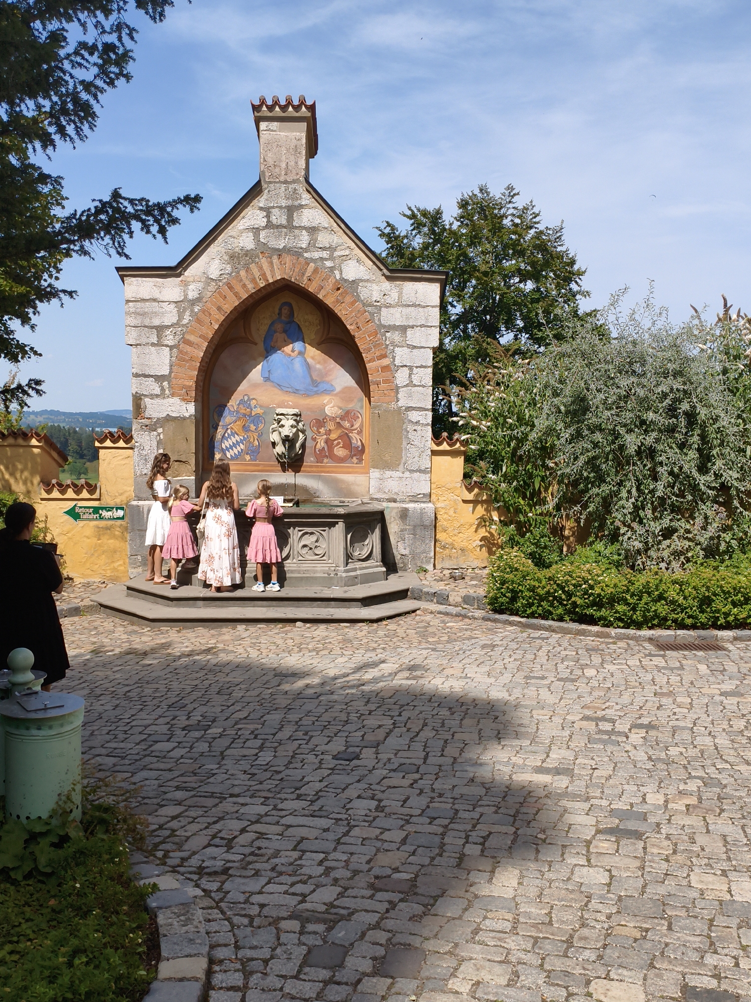 The image depicts a religious fountain structure located in Schwangau, Germany, outside of Neuschwanstein Castle. The structure is adorned with a fresco of the Virgin Mary holding the infant Jesus, surrounded by heraldic symbols and a lion's head spigot. Four young girls, dressed in pink dresses and one in a white dress, are gathered around the fountain, appearing to interact with it. A woman in a black dress stands to the left, observing the scene. The fountain is situated in a cobblestone courtyard, with lush green trees and bushes forming the backdrop. The sky is a clear blue with a few wispy clouds.