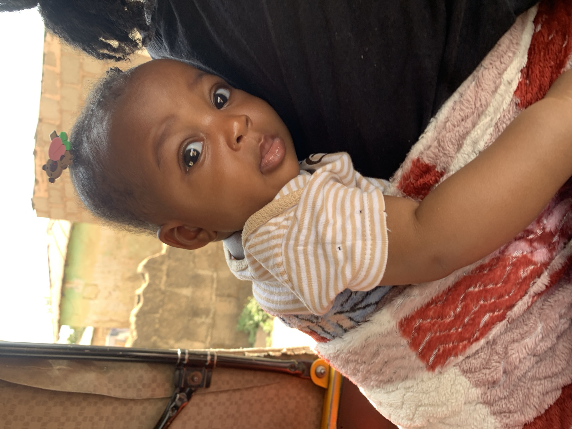A baby with dark skin is held in someone's arms. The baby wears a cream-colored striped shirt and has a brown teddy bear clip in their hair. The baby is wrapped in a multicolored, soft blanket with shades of red, white, and grey. The adult holding the baby is wearing a black shirt. In the background, there is a building, and part of a stroller or carriage is visible in the lower left corner. Based on the available information, pinpointing a precise location within Jos, Nigeria is not possible.