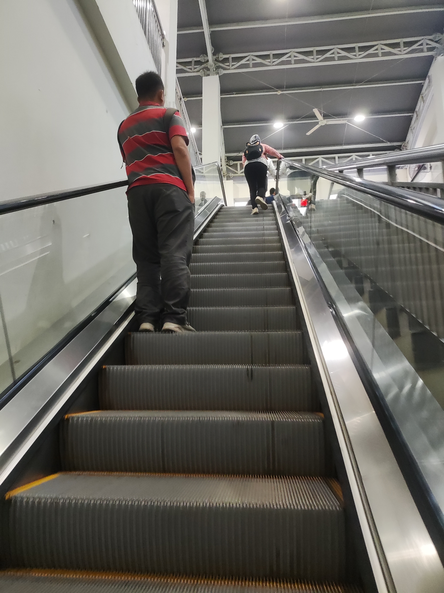 In Telagamurni, Indonesia, an image captures an escalator in operation. A man, wearing a red and grey striped shirt and dark pants, stands on the escalator steps, facing away from the camera. Behind him and further up the escalator, a person wearing a light pink top, dark pants, and a headscarf is also ascending. The escalator is made of dark grey metal with yellow safety lines on the edges of the steps. The surrounding environment appears to be an indoor public space with a high ceiling, exposed metal beams, and lighting fixtures. A ceiling fan is visible in the background. The escalator is flanked by a white wall on the left and a glass railing on the right.