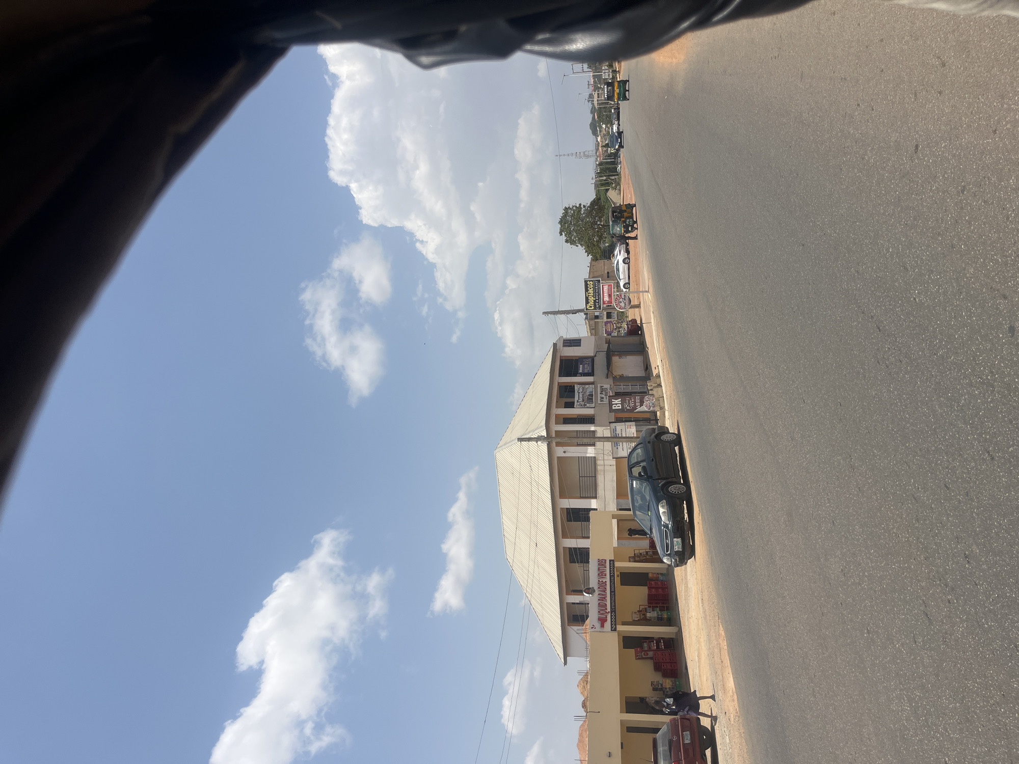 The image captures an urban scene in Jos, Nigeria, as viewed from a moving vehicle. The sky is blue with scattered white clouds. A paved road occupies a significant portion of the frame, leading into the distance. Buildings line the roadside. One notable structure, identifiable as "Cloud Paradise Ventures," appears to be a multi-story building with a light-colored facade. A black car is parked near this building. People are visible near the structures. Other vehicles can be seen further down the road. The perspective is from inside a vehicle, as suggested by a partial view of the vehicle's interior at the top of the frame.
