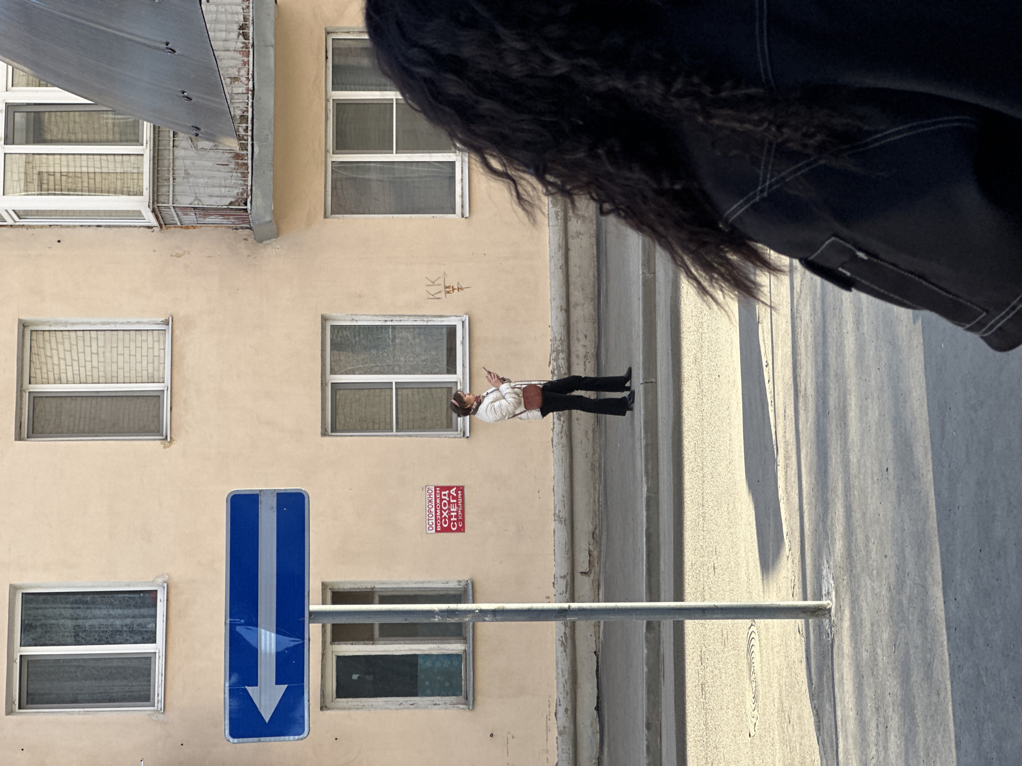 The image, taken in Kazan, Russia, captures an outdoor urban scene. In the foreground, a person with dark, curly hair and wearing a black jacket obscures a portion of the view on the right. The background is dominated by a pale yellow building facade with multiple windows arranged in a grid pattern. A blue traffic sign with a white arrow pointing downwards is visible. A pedestrian is walking along the sidewalk, wearing a light-colored jacket, dark pants, and carrying a brown shoulder bag. A red sign with Cyrillic text is also attached to the building. The scene is lit by sunlight, creating shadows on the sidewalk.