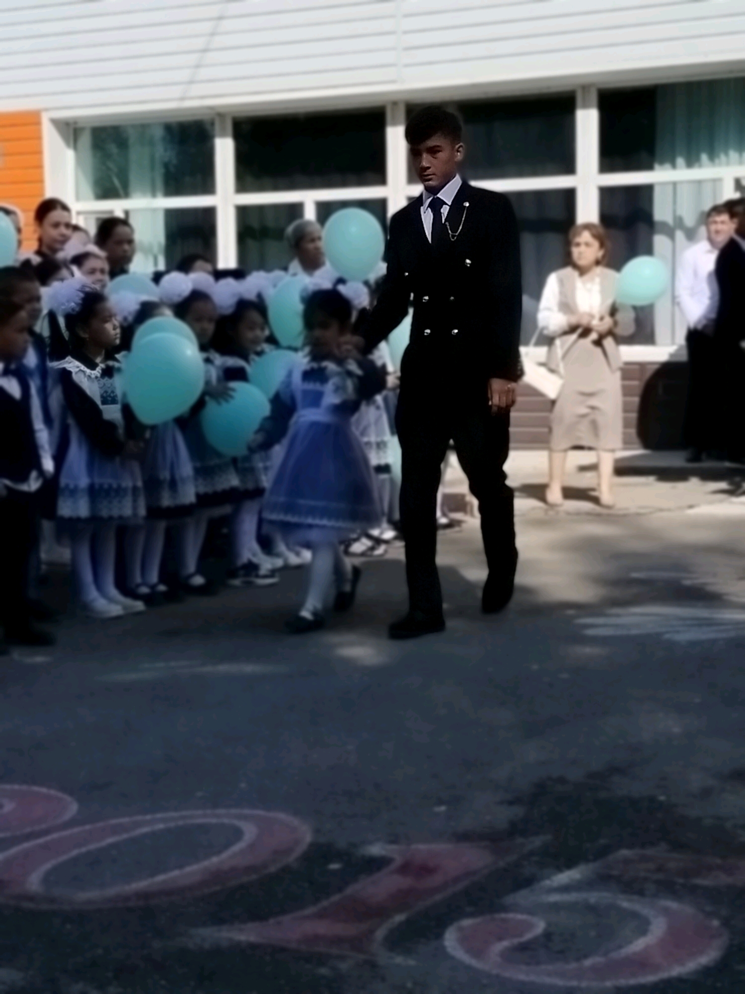 A medium shot shows a group of children, primarily young girls in school uniforms, standing in front of a building. A young man in a black suit with a tie and a waistcoat is walking past them. The girls are holding light blue balloons. The building appears to be a school, with large windows. The ground in front of the building has chalk markings, possibly numbers or letters. The overall scene suggests a school event, possibly a celebration or the first day of school. The presence of chalk markings on the ground hints at a possible location within a city, and given the context, it could be a public space like a schoolyard in Көлкент, Kazakhstan.