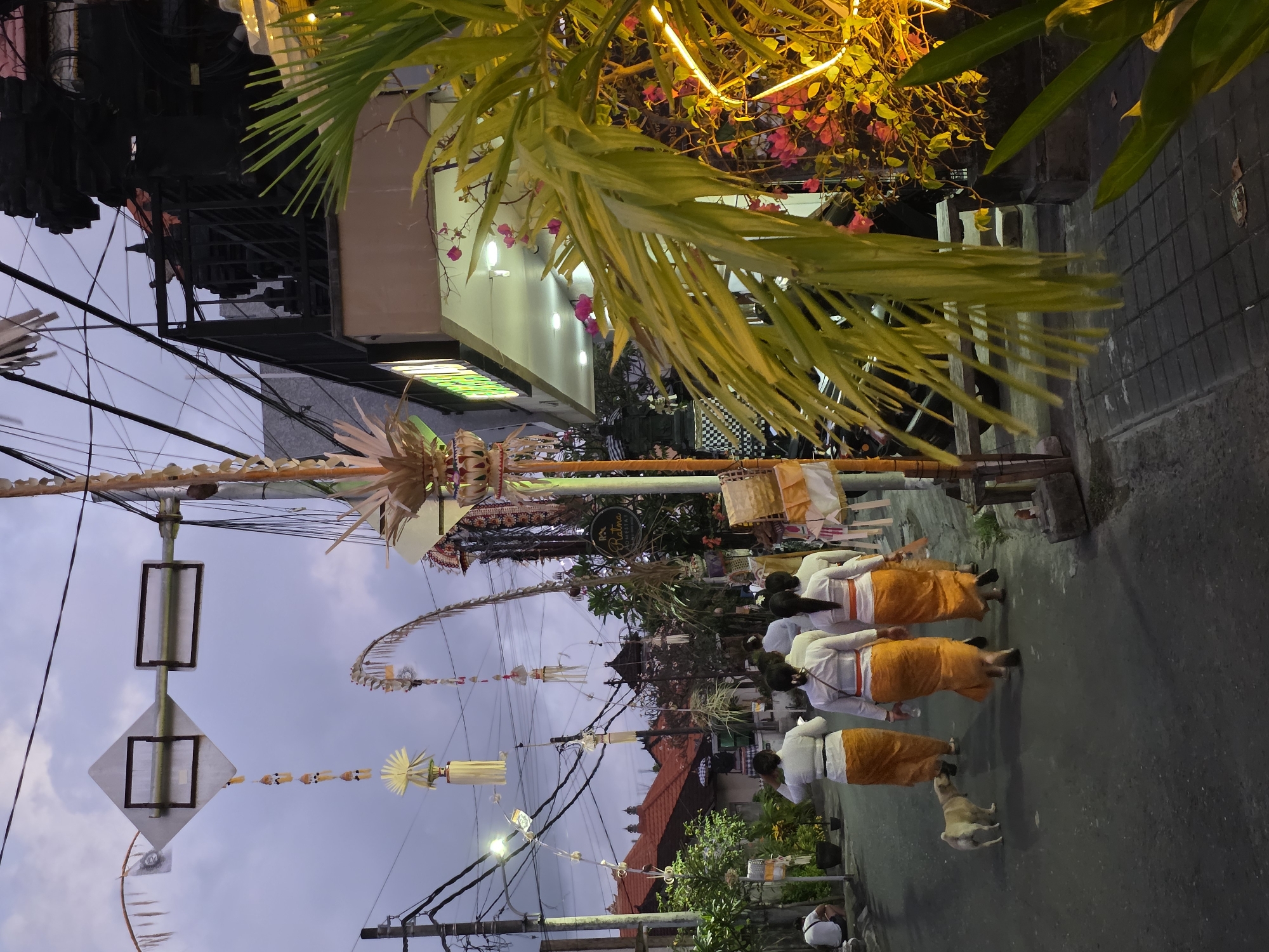 In North Kuta, Indonesia, as dusk settles, a group of women in traditional white tops and orange sarongs walk down a street, accompanied by a small tan dog. The street is lined with buildings adorned with decorative offerings, including tall woven structures and hanging ornaments. Overhead, power lines crisscross the sky, illuminated by streetlights. A street sign is visible above, and a palm frond with yellow lights provides a vibrant foreground element. The atmosphere suggests a cultural or religious procession.