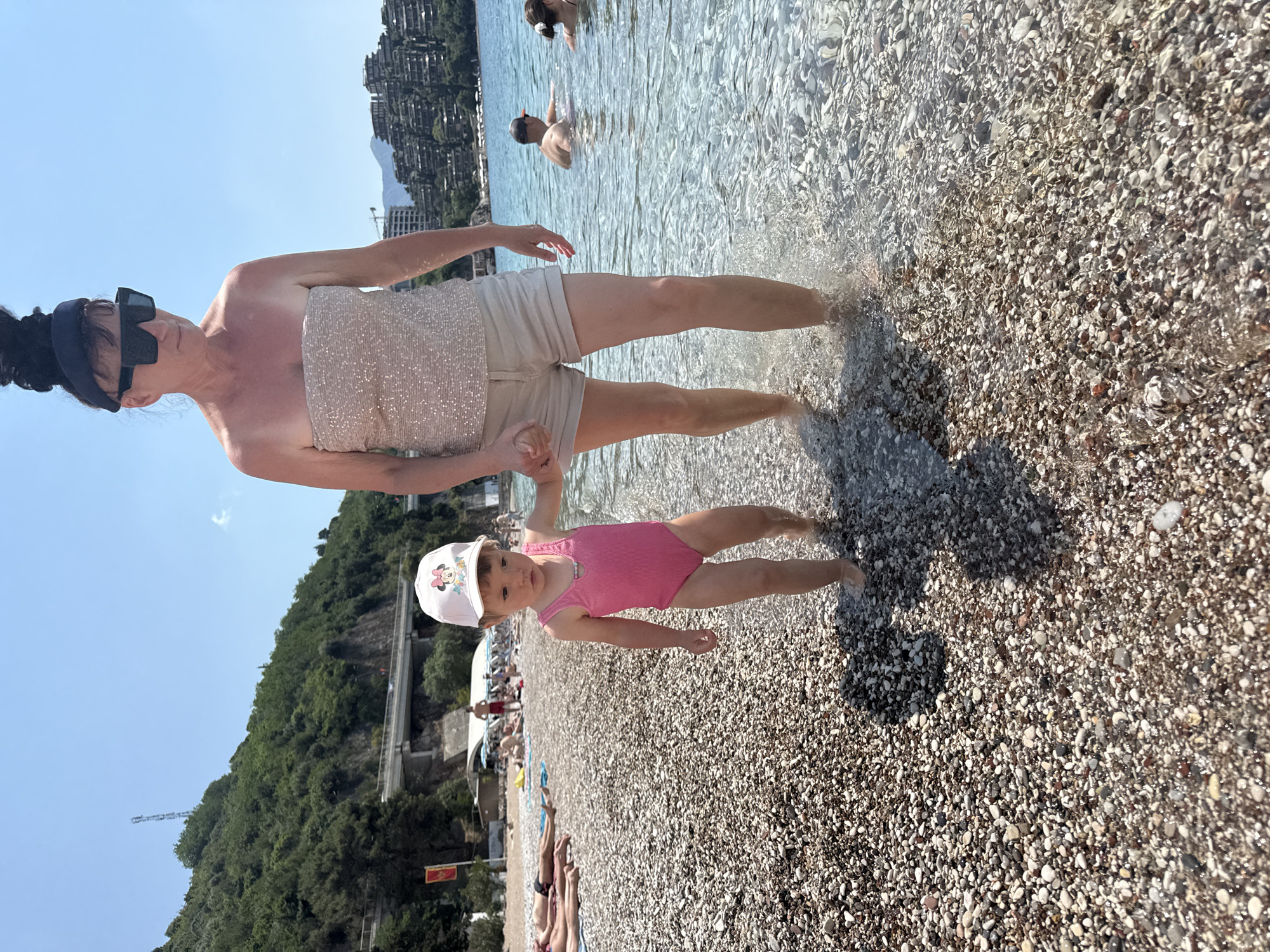 The image depicts a woman and a young girl standing in the shallow water at a beach. The beach is pebbly, with various sizes and colors of pebbles visible. In the background, there's a shoreline with buildings and vegetation, indicating an urbanized area. The water is clear enough to see the pebbles beneath its surface. The woman is wearing a strapless sequined top and beige shorts. She's also wearing sunglasses and a dark headscarf. She is holding the girl's hand. The girl is wearing a pink swimsuit and a white hat. People are swimming in the background.
The location is the city of Budva, Montenegro, as indicated by the typical landscape and architecture of the area. The image captures a moment of interaction between the woman and the girl, likely enjoying a day at the beach.