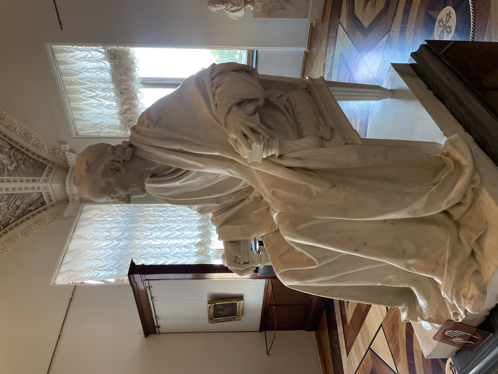 Here is a description of the image:
The image shows a sculpture of a man seated and draped in a robe. He appears to be an older male with a thoughtful expression. He's holding a book or tablet. The sculpture is situated in what appears to be a museum or historical building, given the ornate ceiling and patterned wooden floor. There are cabinets and a framed picture visible in the background, along with a window covered by a patterned curtain. A plaque on the base of the sculpture contains a logo depicting a "Do Not Touch" sign.