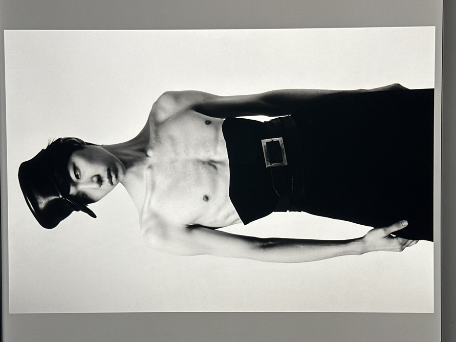 A monochromatic image features a slender, androgynous figure with a hat, torso exposed. The subject wears dark trousers cinched with a belt featuring a rectangular buckle. The individual poses against a white backdrop, exhibiting a tilted head and one arm extended with the hand partially visible. The lighting emphasizes shadows across the chest and arms, accentuating the figure's contours. The composition suggests a fashion editorial or artistic portraiture style.