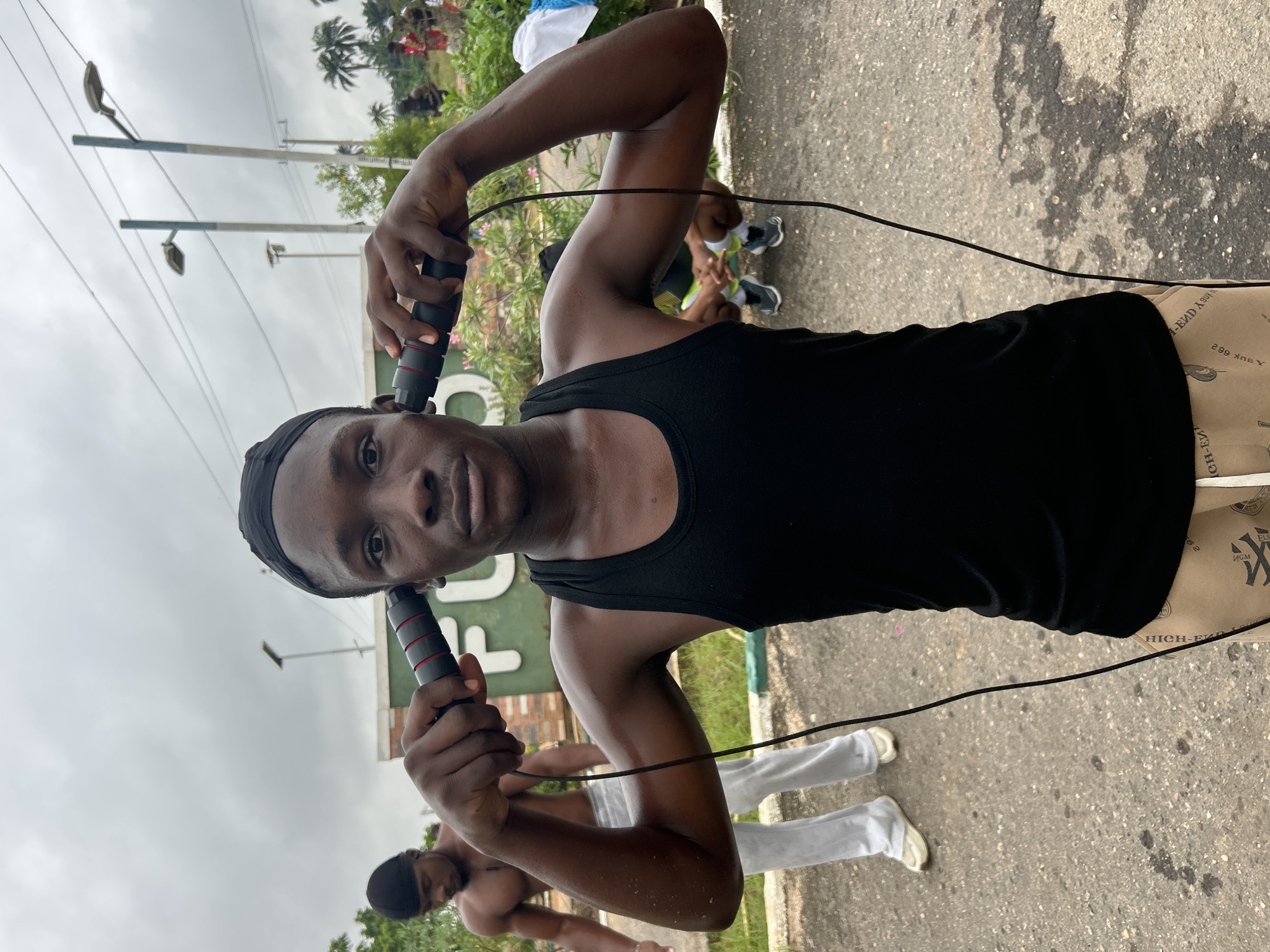 In the city of Ihiagwa, Nigeria, a young Black male is captured in a medium shot, holding a jump rope horizontally in front of his head, with the handles pressed against his ears. He wears a black tank top and tan pants with a repeated pattern of text and symbols. His gaze is directed forward, his expression neutral. To his left, another Black male, shirtless and wearing a black durag, stands with his head down. In the background, a large green sign with white lettering, partially obscured by foliage, is visible. Two lampposts stand tall against an overcast sky. The scene appears to be an outdoor area, possibly a recreational space, with pavement underfoot and greenery surrounding the area.