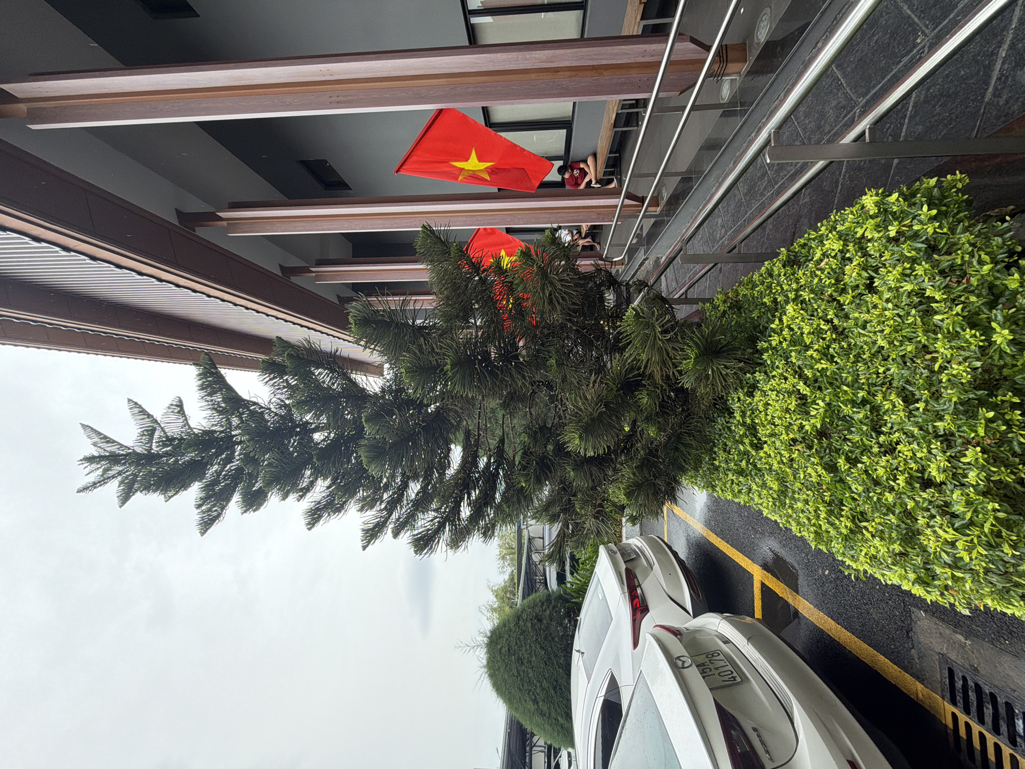 In Đà Nẵng, Vietnam, a parking lot adjacent to a modern building features a mature evergreen tree. Two white cars are parked, one with license plate "15A 40178". A manicured hedge runs along the edge of the parking lot. The building's exterior is characterized by wooden columns and a dark gray facade. Two red Vietnamese flags with yellow stars are displayed, one prominently in the foreground. A person is seated on a wooden bench under an overhang of the building, appearing to be looking down. The sky is overcast.