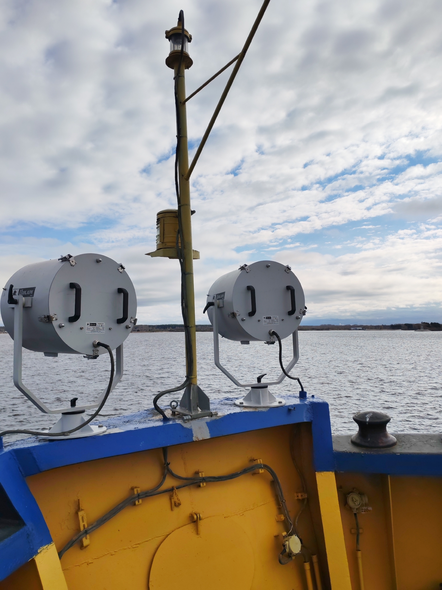 The image shows the deck of a vessel, possibly a tugboat or small ship, likely docked in Rybinsk, Russia, given the industrial appearance of the landscape in the background and the Cyrillic text present on one of the objects.
The vessel's deck is painted yellow with blue trim. There are two cylindrical gray lights, possibly searchlights, mounted on white bases. The lights have handles on their sides. A vertical yellow pole is centrally located between the lights, topped with a navigational light fixture.
Electrical cables run along the interior of the vessel's deck. A black bollard is visible on the right side of the image. The background shows water and a distant shoreline with trees and some industrial structures. The sky is overcast with clouds.