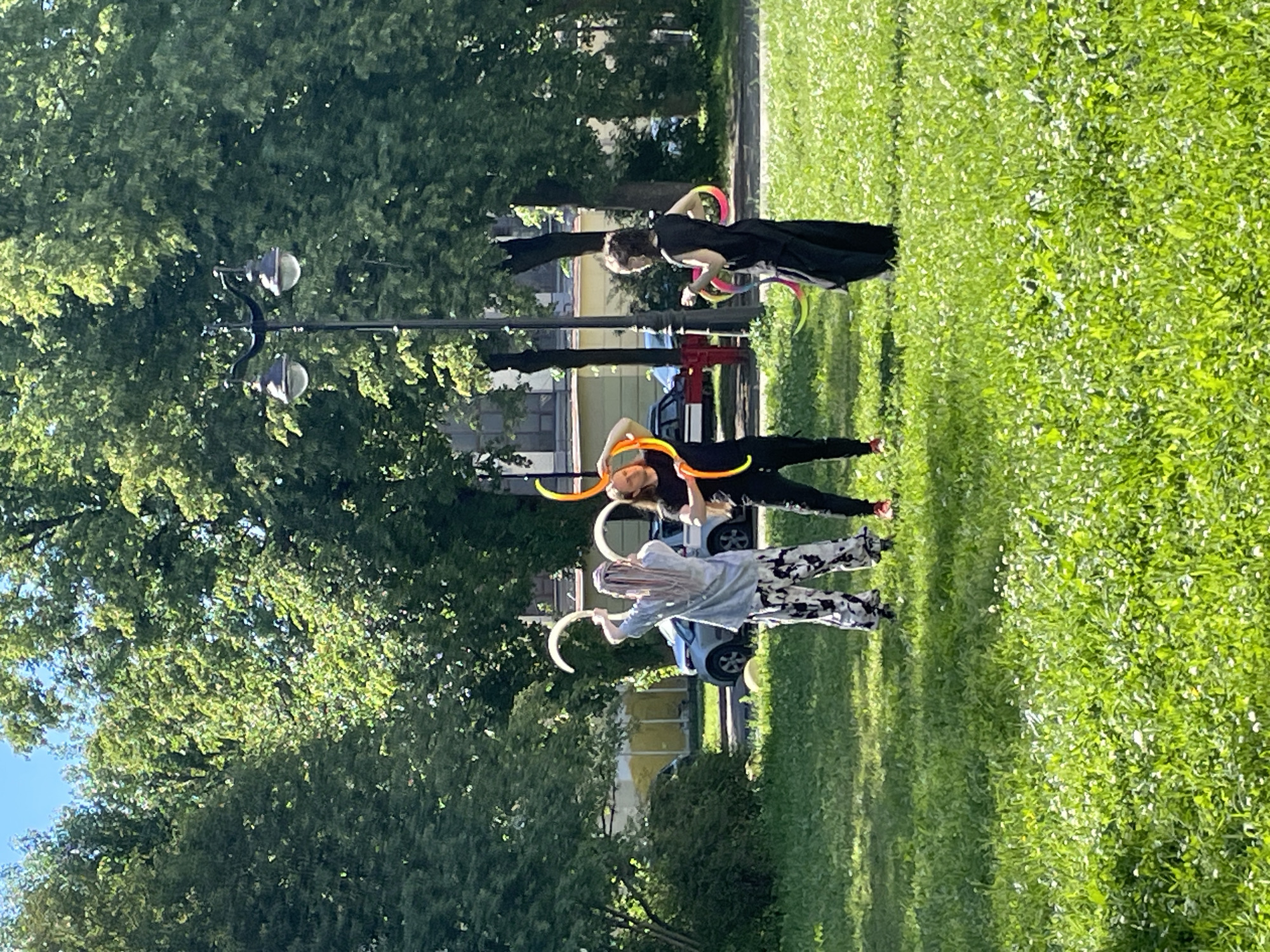 In Saint Petersburg, Russia, three individuals are captured mid-hoop dance in a grassy park setting. The sun shines brightly, casting long shadows and illuminating the lush green grass. Trees provide a natural backdrop, with the hint of buildings and streetlights visible in the distance.
The subjects are dressed in casual attire. The person in the foreground, wearing a light-colored shirt and patterned pants, is holding two white hoops. Behind them, another individual in a black outfit is skillfully manipulating multiple colorful hoops around their body. The third person, also in dark clothing, is similarly engaged with several hoops. The overall atmosphere suggests a practice session or a performance, with the individuals focused on their movements and the colorful hoops adding a dynamic visual element to the scene. The time of day appears to be late afternoon, given the quality of the light and shadows.