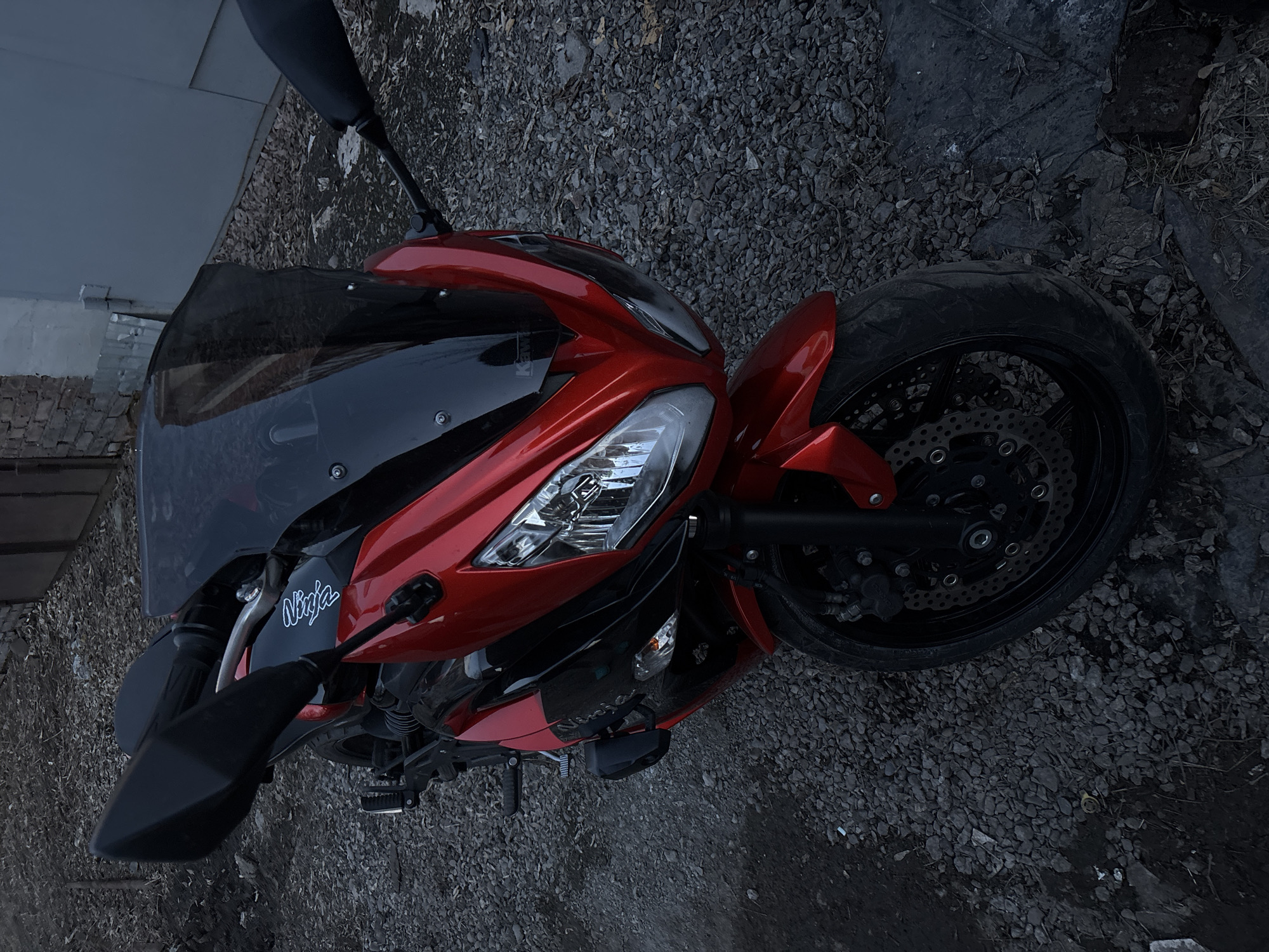 The image shows a red and black Kawasaki Ninja motorcycle parked on a gravel surface. The front wheel is prominently visible with its disc brakes. The bike's headlight, windscreen, and rearview mirrors are also in view. The surroundings consist of gravel and a building with a brick foundation and a gray facade, potentially located in an urban area. Based on the prompt constraints, there is no information to determine the specific location in the city of Krasnoyarsk, Russia.