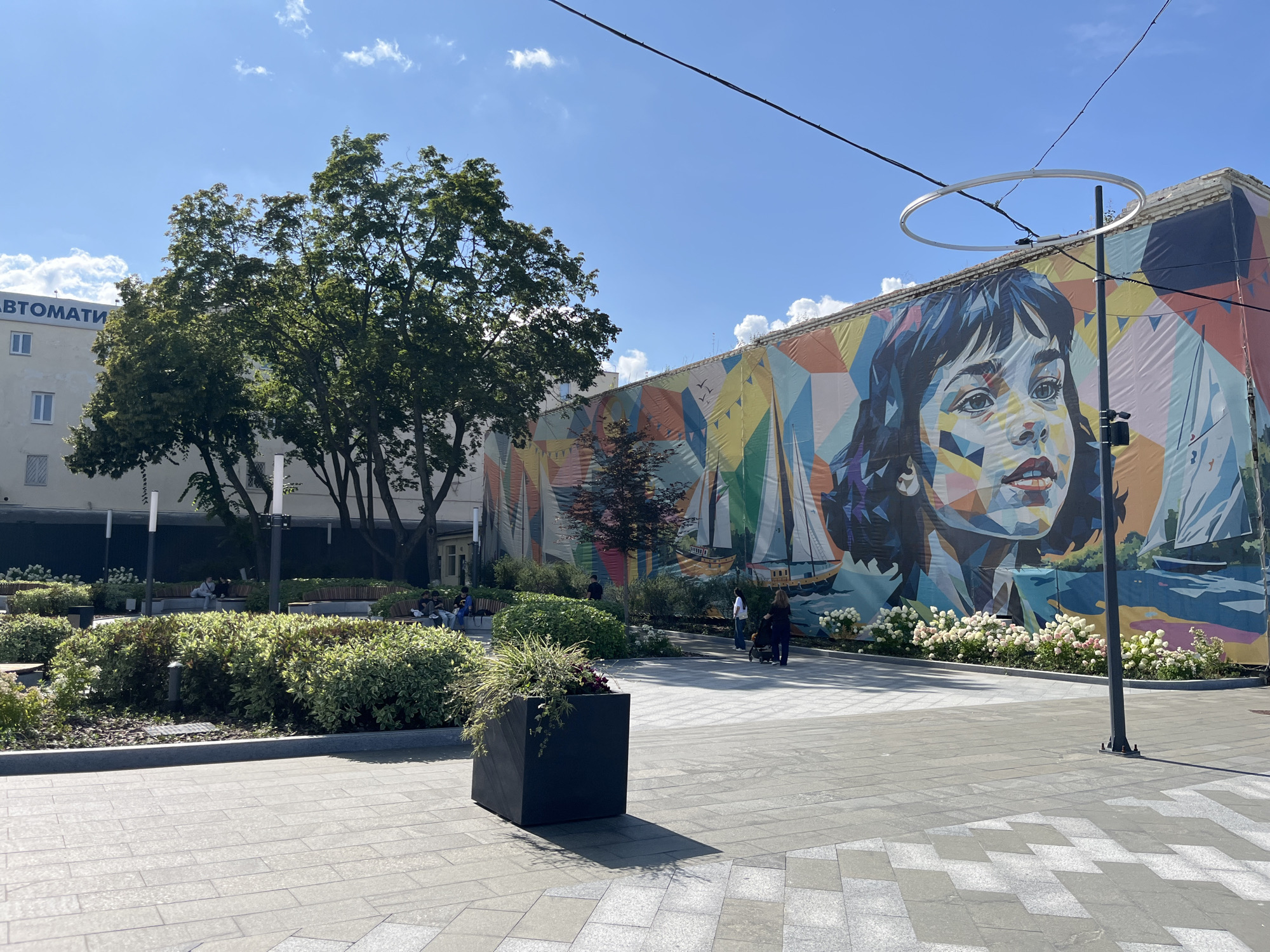 The image depicts a sunny day in a public plaza in Kharkiv, Ukraine, likely near the Kharkiv National University of Municipal Economy. A large, vibrant mural adorns the side of a building, featuring a stylized portrait of a young woman and sailboats against a geometric, colorful backdrop. The mural covers a significant portion of the building's facade. In the foreground, a paved plaza is visible, with large, square gray tiles and a geometric pattern of lighter gray tiles creating a visual effect. A large, dark square planter box with greenery sits in the middle ground to the left. Beyond the planter, a well-maintained garden area with bushes and trees is present. Benches with people seated are visible in the shaded area beneath the trees. Several individuals are walking through the plaza, including a person pushing a stroller. Power lines and a circular light fixture are suspended above the scene, with the clear blue sky and scattered clouds overhead.