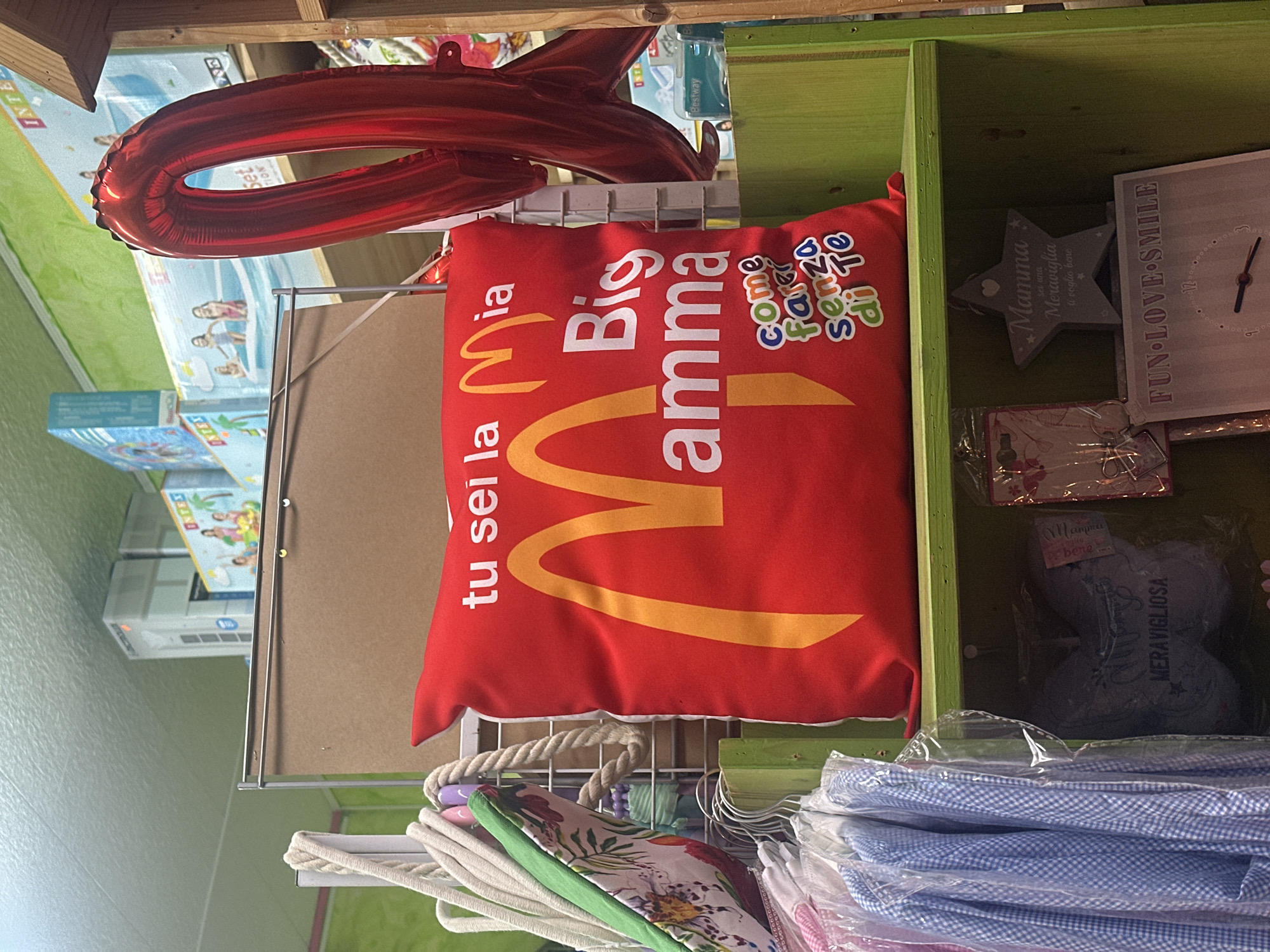 This image captures an interior scene from a gift or novelty shop, likely located in Lipari, Italy. The main subject is a vibrant red cushion, prominently displayed on a white wire rack. The cushion features a large, golden, McDonald's-style "M" logo and Italian text that reads, "tu sei la Mia Big Mamma come farei senza di te," which translates to "you are my Big Mom, how would I do without you."
Above the cushion, a large, glossy red foil balloon, possibly in the shape of a number or letter, is partially visible. To the right of the cushion, various gift items are arranged within green wooden shelves. These include a grey star-shaped plaque with the Italian inscription "Mamma Meraviglia ti voglio bene" (Wonderful Mom, I love you), and a white plaque or clock that reads "FUN LOVE SMILE." Below these, a blue heart-shaped item in plastic packaging is visible, marked with "MERAVIGLIOSA" (Wonderful).
Below the red cushion, several textile items are stacked or hanging, including a coiled rope, fabric with a floral pattern, and neatly folded blue checkered cloth, suggesting various household or personal items for sale. In the background, out of focus, product boxes for brands like "Intex" and "Bestway" can be seen, indicating the shop may also sell leisure or outdoor equipment. The artificial lighting suggests an indoor setting. The items on display point to a retail environment specializing in affectionate gifts and souvenirs, suitable for family members, particularly mothers.