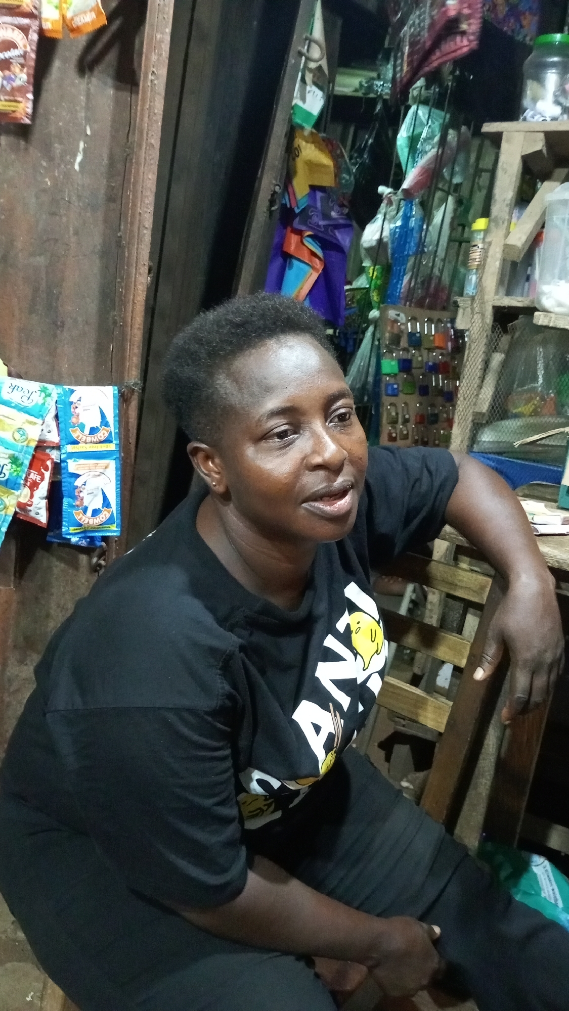 The image features a woman with dark skin and short, textured hair, seated in what appears to be a small shop or stall. She is wearing a black short-sleeved T-shirt with a partially visible graphic design on the front, which includes a yellow chick-like character and white text that begins with "AN-". She is also wearing dark pants. Her left arm rests on the back of a wooden chair, and her right hand is on her leg. She is looking towards the right side of the frame with a slightly open mouth and a neutral, contemplative expression.
The background is filled with various items commonly found in a local store. To her left, several sachets of products are hanging, including "COWBELL" and "PEAK" (likely milk powder) and "NESCAFE" (coffee). The wall behind her appears to be made of wooden planks. To her right, a wooden shelving unit displays an array of colorful padlocks hanging on hooks, along with other general merchandise such as plastic containers and rolled umbrellas. The lighting is somewhat dim, indicating an indoor setting. The scene suggests an ordinary moment in a busy market or convenience store in Onitsha, Nigeria.