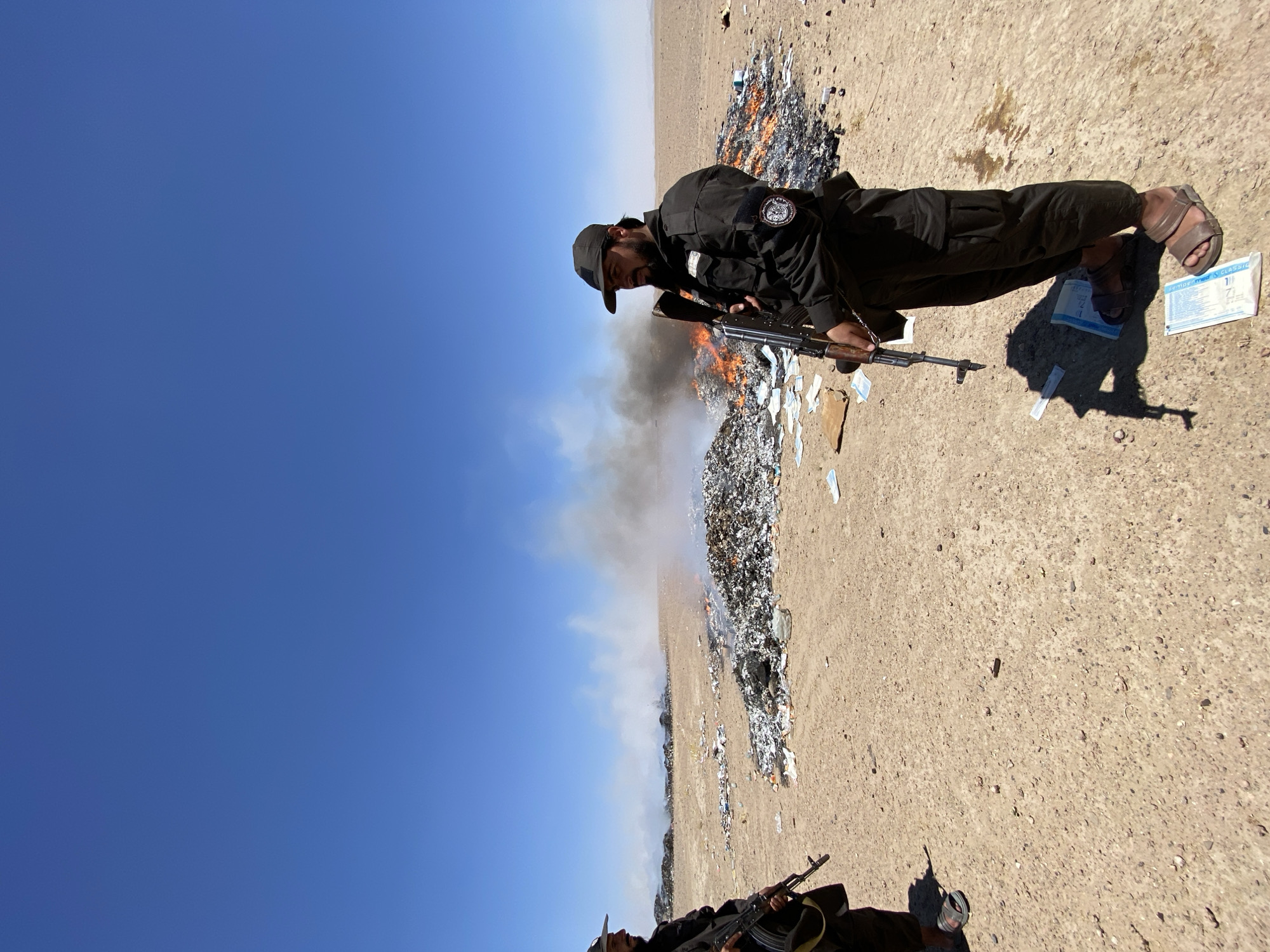 In Mandisar, Afghanistan, two men, likely security personnel based on their dark uniforms and tactical vests, stand guard. One man, in the foreground, holds an assault rifle and wears a baseball cap. The other, further in the background, also carries a rifle. They are positioned in a desert landscape under a clear blue sky. In the mid-ground, a large pile of miscellaneous items, appearing to be trash and possibly some documents or paper, is engulfed in flames and producing thick smoke. The ground around the fire is strewn with debris.