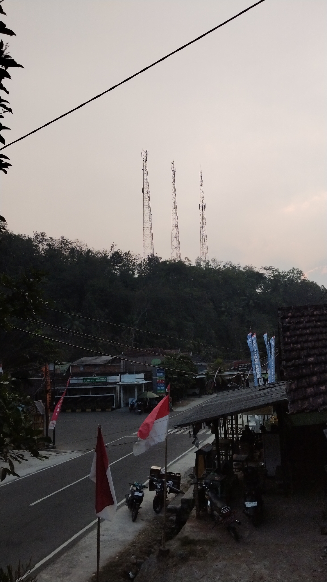 The image depicts a street scene in the city of Tegalombo, Indonesia. In the background, three tall communication towers stand on a tree-covered hill. The foreground features a road with two Indonesian flags planted on the side. Several motorcycles are parked along the road and in front of a small establishment with a covered seating area. Buildings with signs, one reading "TOILET UMUM" (public toilet), are visible on the opposite side of the road. The sky is overcast, suggesting it might be dusk or early morning. The overall impression is that of a quiet, rural roadside with a few functional structures and signs of daily life.